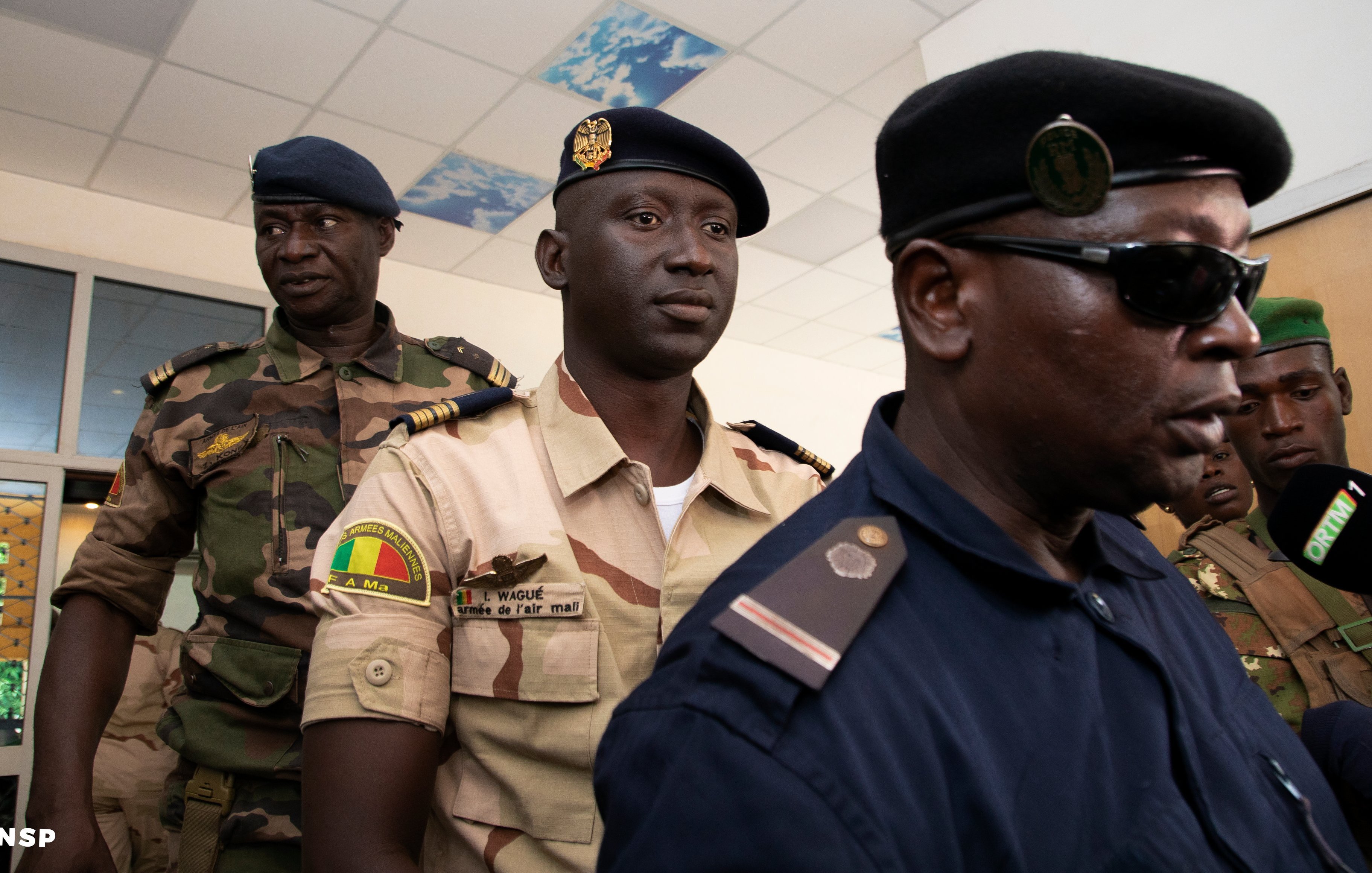 L'image montre un groupe d'hommes en uniforme, probablement des membres des forces de sécurité. Au premier plan, on peut voir un homme en uniforme sombre, portant un chapeau, qui parle à un microphone. Il semble être en train de faire une déclaration ou une interview. À l'arrière-plan, deux autres hommes en uniforme sont présents. L'un d'eux porte un uniforme plus clair avec des insignes, tandis que l'autre a un uniforme de camouflage. L'environnement semble être un bâtiment officiel, avec des murs clairs et un plafond décoré. L'atmosphère peut être perçue comme sérieuse et formelle.