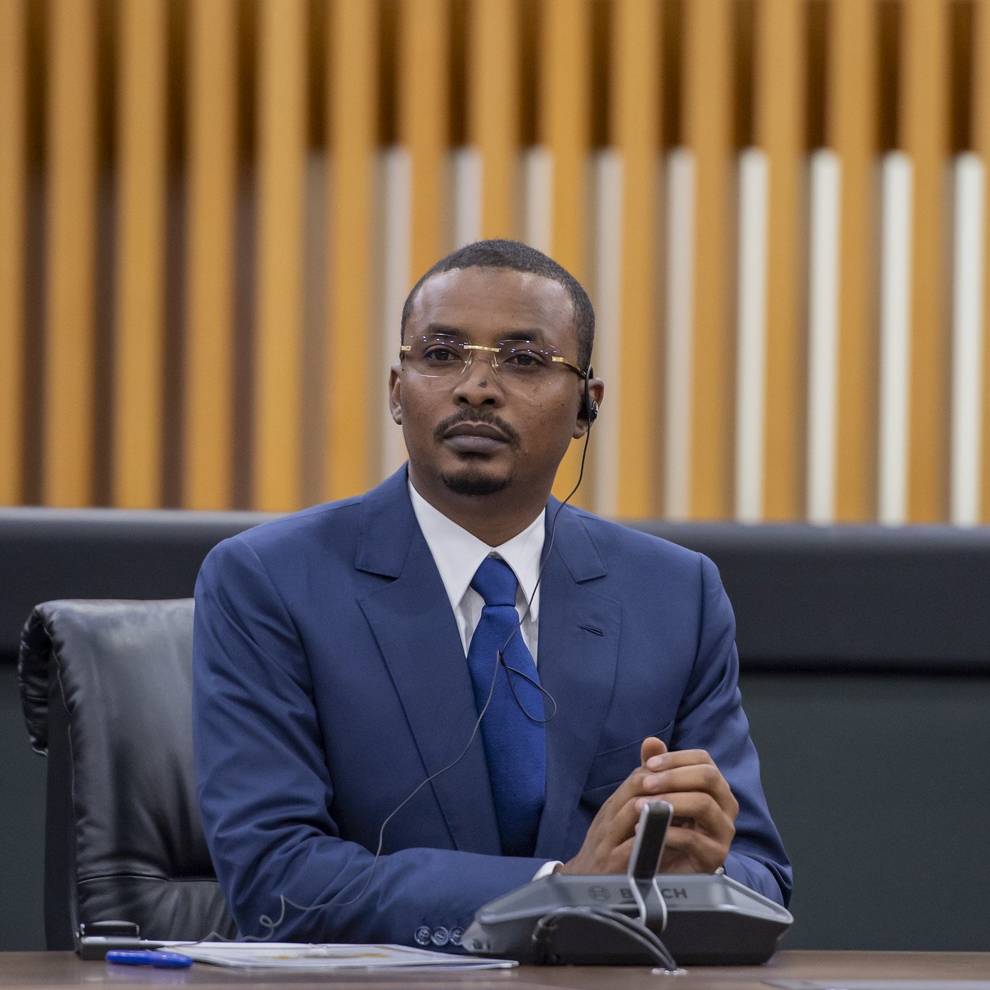 Dans cette image, un homme est assis à une table dans un environnement formel, probablement lors d'une réunion ou d'une conférence. Il porte un costume bleu, une chemise blanche et une cravate bleue. Ses cheveux sont courts et bien coiffés. L'homme semble concentré et attentif, avec un léger froncement de sourcils et les mains croisées devant lui. À son oreille, on peut voir un écouteur, ce qui suggère qu'il écoute probablement une traduction ou une intervention en cours. En arrière-plan, on distingue des panneaux en bois aux lignes verticales, ajoutant une touche moderne à l'espace. L'atmosphère générale est professionnelle et sérieuse.