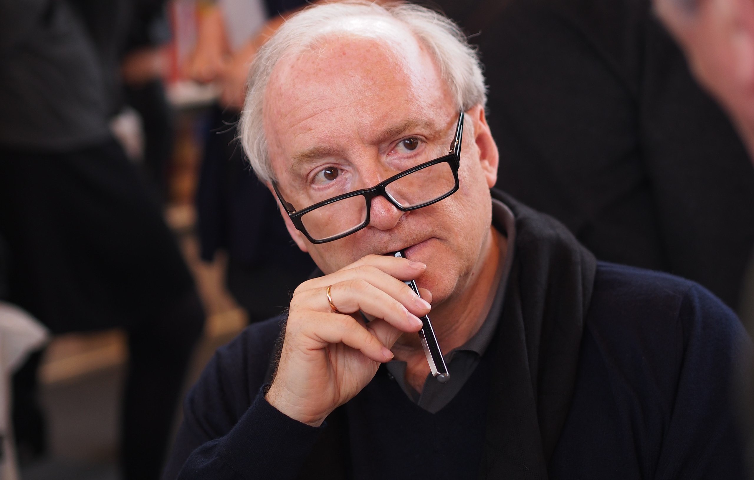 L'image montre un homme assis, vêtu d'un pull sombre, avec des lunettes sur le nez. Il porte une expression pensive, tenant un stylo près de sa bouche, comme s'il était en train de réfléchir ou de considérer quelque chose de manière approfondie. En arrière-plan, on distingue d'autres personnes floues, suggérant une ambiance de discussion ou d'échange, typique d'un événement de rencontre ou de signature de livres. La lumière est douce, créant une atmosphère chaleureuse.