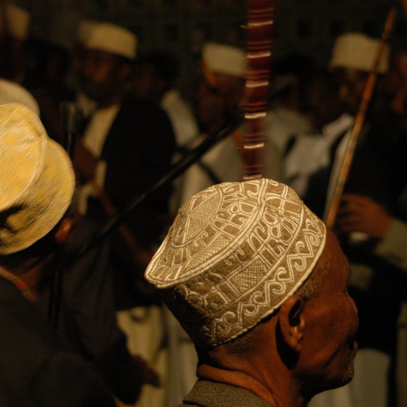 L'image présente un groupe de personnes, principalement des hommes, de profil. Ils portent des chapeaux traditionnels, souvent ornés de motifs délicats, qui varient en couleurs et en designs. En arrière-plan, on peut distinguer d'autres figures et instruments de musique, suggérant une ambiance festive ou rituelle. La lumière est tamisée, créant une atmosphère intime. Les visages des hommes sont flous, mais leur posture et leurs chapeaux ajoutent une touche culturelle à la scène.