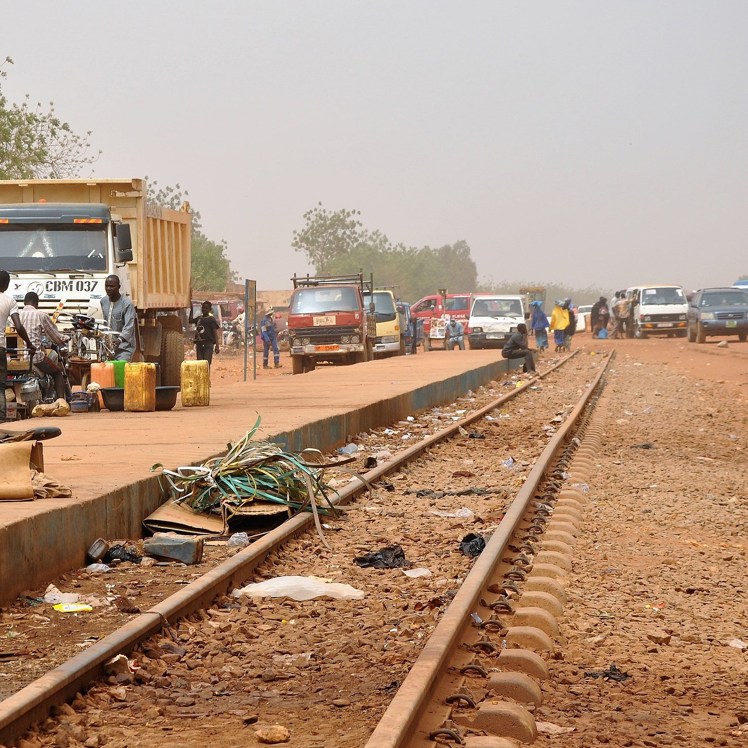L'image dépeint une scène animée le long d'une voie ferrée. Sur la droite, des rails en métal s'étendent, entourés de poussière. À proximité, on aperçoit plusieurs véhicules, notamment des camions et des voitures, stationnés ou en mouvement. Des personnes se regroupent sur le côté, certains discutant, d'autres transportant des marchandises. Sur le sol, on peut distinguer divers objets laissés là, comme des déchets, des morceaux de métal et des câbles. À gauche, une petite charrette à deux roues est partiellement visible. En arrière-plan, la végétation est clairsemée, avec quelques arbres. Le ciel est d'un ton brumeux, suggérant une chaleur intense et un environnement rural. L'atmosphère générale est à la fois occupée et rustique, évoquant l'activité quotidienne dans une région où la vie s'organise autour des transports.