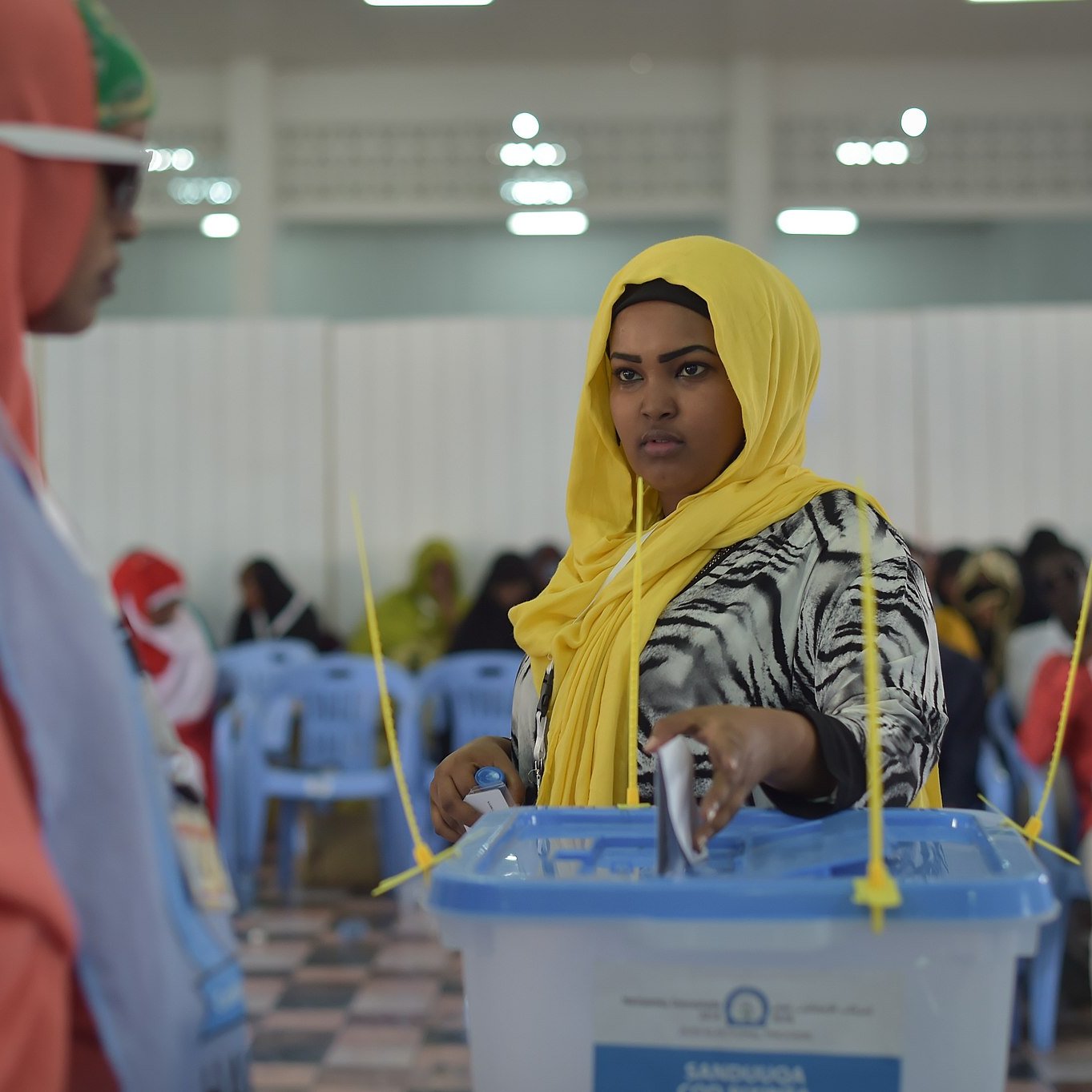 L'image montre une scène de vote dans un lieu organisé. À gauche, une femme porte un hijab orange et des lunettes de soleil, regardant vers une autre femme, qui est située à droite. Cette dernière, en hijab jaune, semble concentrée alors qu'elle dépose son bulletin dans une urne transparente devant elle. L'intérieur de la salle est éclairé, avec des chaises en plastique bleues disposées en rangées. On peut percevoir une atmosphère de participation et d'engagement civique. Des femmes en arrière-plan sont également présentes, créant une ambiance communautaire.