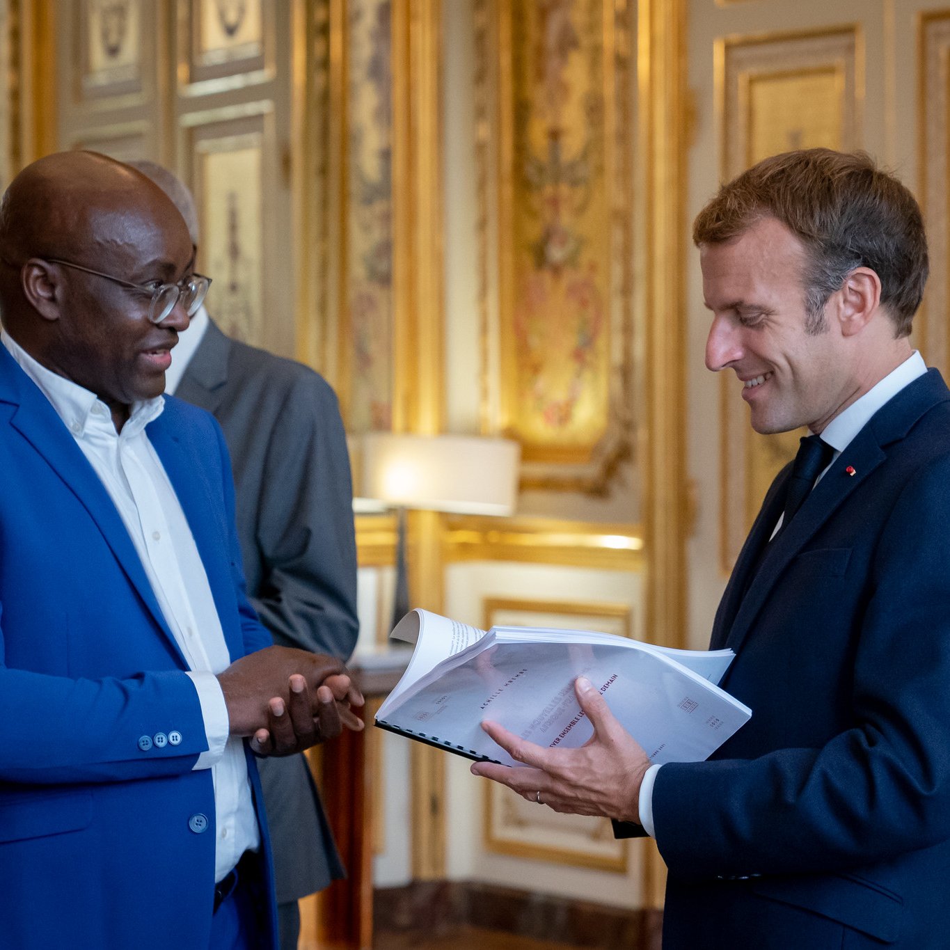 L'image montre deux hommes se tenant debout dans une pièce élégante et ornée. L'un des hommes porte un costume bleu vif et sourit tandis qu'il parle. L'autre homme, vêtu d'un costume sombre, tient des documents dans ses mains et lui répond avec un léger sourire. La décoration de la pièce est luxueuse, avec des murs dorés et des éléments classiques qui évoquent un cadre formel et historique. Il y a une lumière chaleureuse, créant une ambiance conviviale à cet échange.