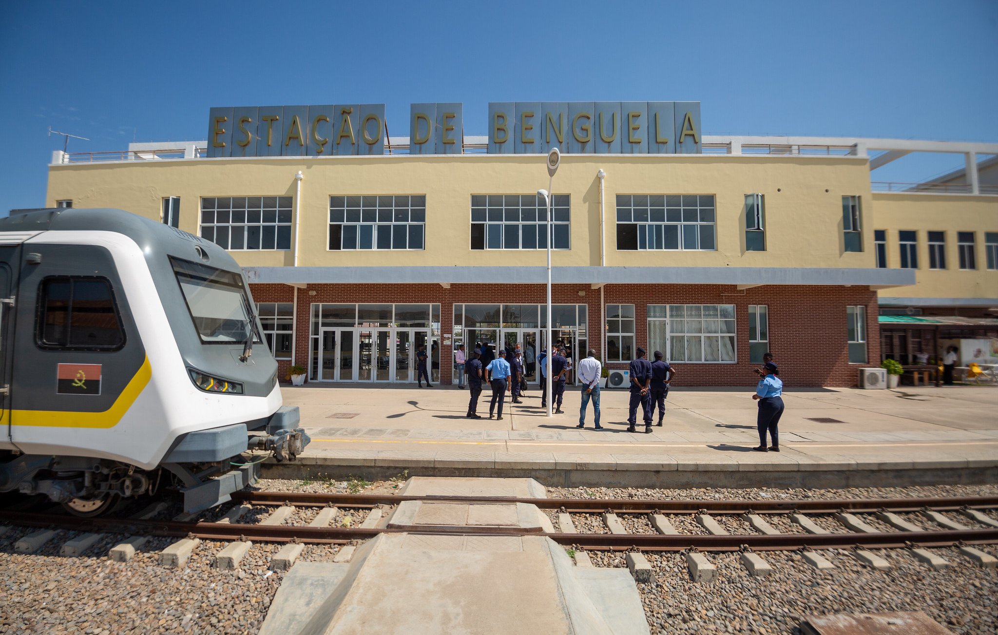 Cette image montre la gare de Benguela, un bâtiment aux couleurs pâles et au style architectural distinctif. À l'avant, on voit un train moderne garé sur une voie ferrée. L'architecture de la gare est caractérisée par des fenêtres spacieuses qui laissent entrer beaucoup de lumière naturelle. Devant la gare, un groupe de personnes, probablement des voyageurs ou du personnel, se tient debout, discutant ou attendant. Le ciel est clair et ensoleillé, créant une ambiance vive et accueillante. Les rails de la gare sont visibles au premier plan, ajoutant au contexte ferroviaire de la scène.