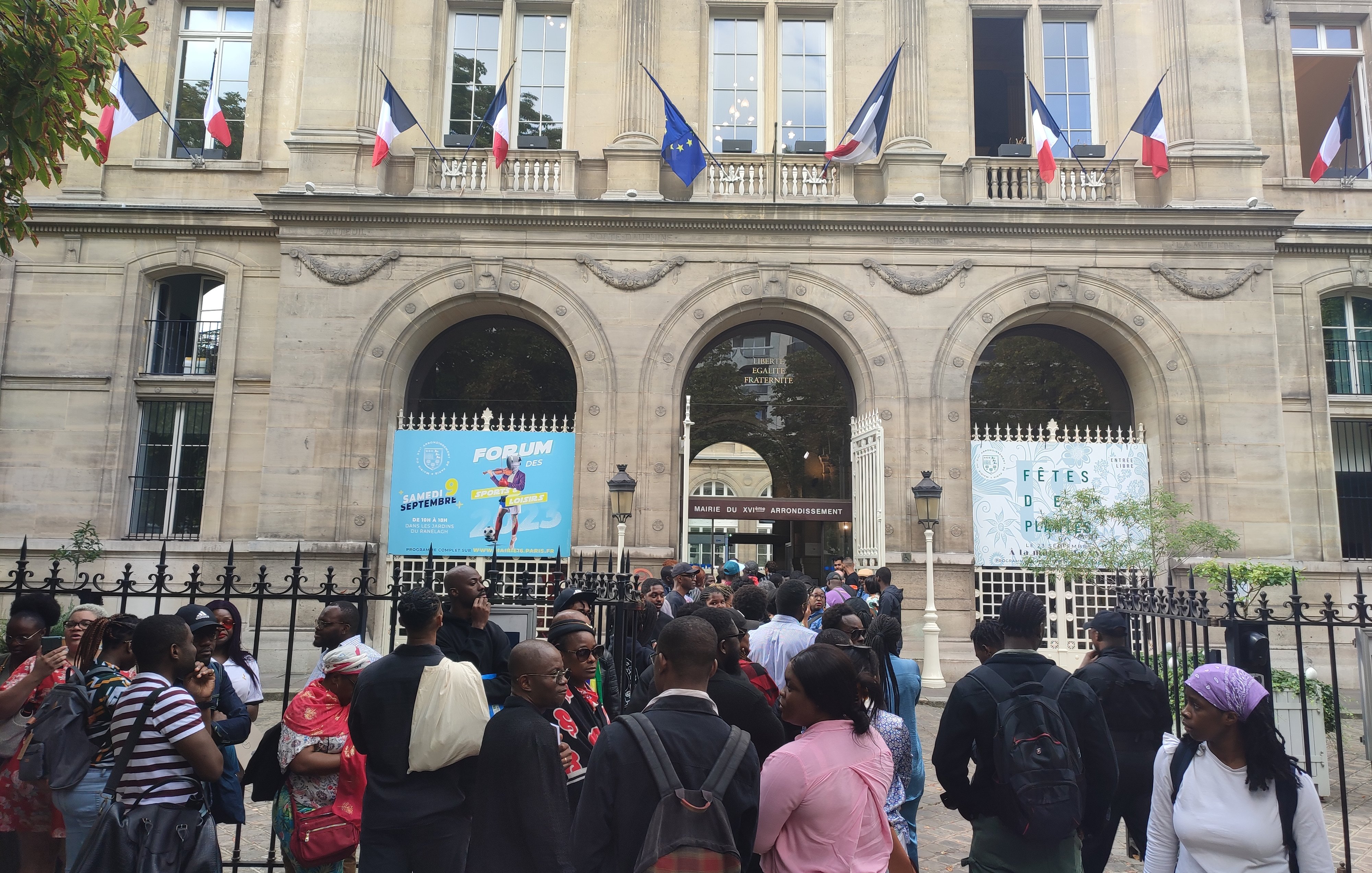 L'image montre un bâtiment imposant avec une architecture classique, visible grâce à ses grandes fenêtres et ses arches. Devant ce bâtiment, un groupe de personnes se rassemble, créant une atmosphère animée. Des drapeaux français flottent au-dessus d'eux, témoignant de l'importance de ce lieu. Les visiteurs semblent variés en termes d'âge et de style vestimentaire, ce qui reflète une diversité culturelle. À l'arrière-plan, des affiches colorées annoncent un événement, ajoutant une touche festive à la scène. L'ensemble dégage une ambiance d'attente et d'excitation.