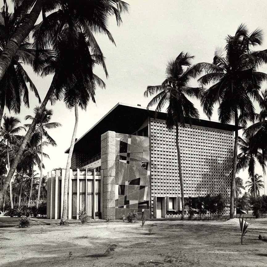 L'image montre un bâtiment moderne, avec une architecture distincte et géométrique. Le mur de la façade est ornée de motifs en relief, créant un jeu d'ombres et de lumière. Des colonnes blanches se dressent devant l'entrée principale, ajoutant une touche de classicisme à la structure. Autour du bâtiment, des palmiers élancés se balancent doucement, donnant une ambiance tropicale. Le sol est nu, avec une végétation assez faible, ce qui permet de mettre en valeur l'architecture. La scène semble baignée dans une lumière naturelle, évoquant une atmosphère calme et sereine.