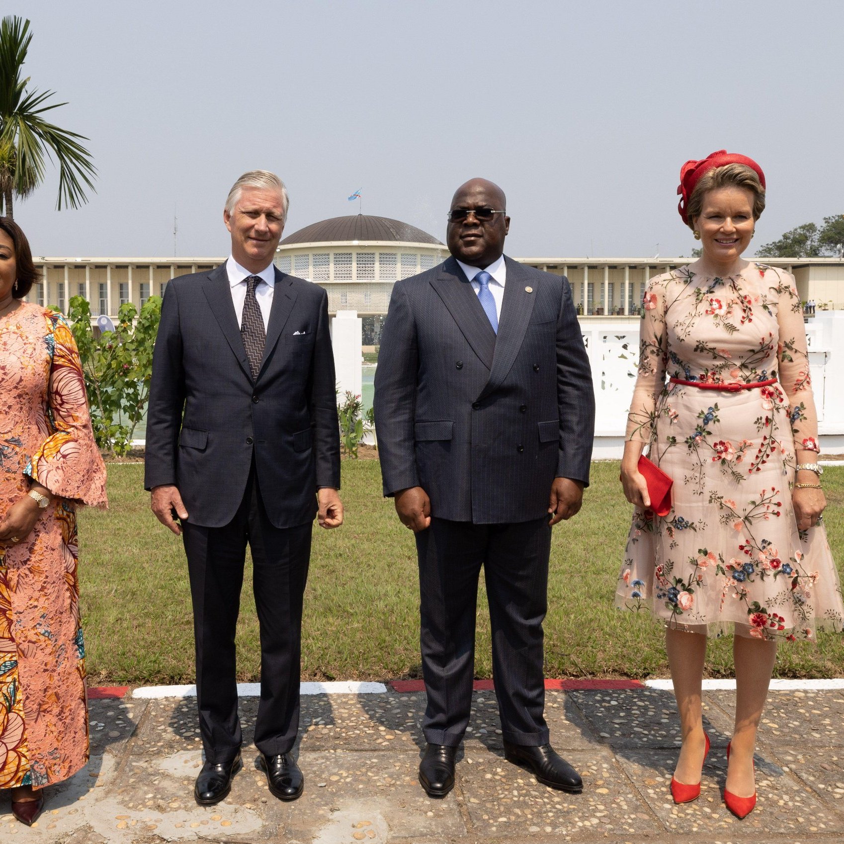 L'image montre un groupe de quatre personnes se tenant debout dans un jardin bien entretenu. À gauche, une femme porte une robe longue à motifs floraux et des manches évasées. À côté d'elle, un homme en costume sombre est debout, affichant une expression chaleureuse. Ensuite, un autre homme, également en costume, a une posture amicale et est au centre de l'image. Finalement, à droite, une femme porte une robe à fleurs avec un joli chapeau assorti et un sac à main rouge, se tenant avec élégance. En arrière-plan, on aperçoit un bâtiment imposant et un palmier qui ajoutent une atmosphère exotique à la scène. Le ciel est clair, ce qui contribue à un sentiment de rencontre officielle.