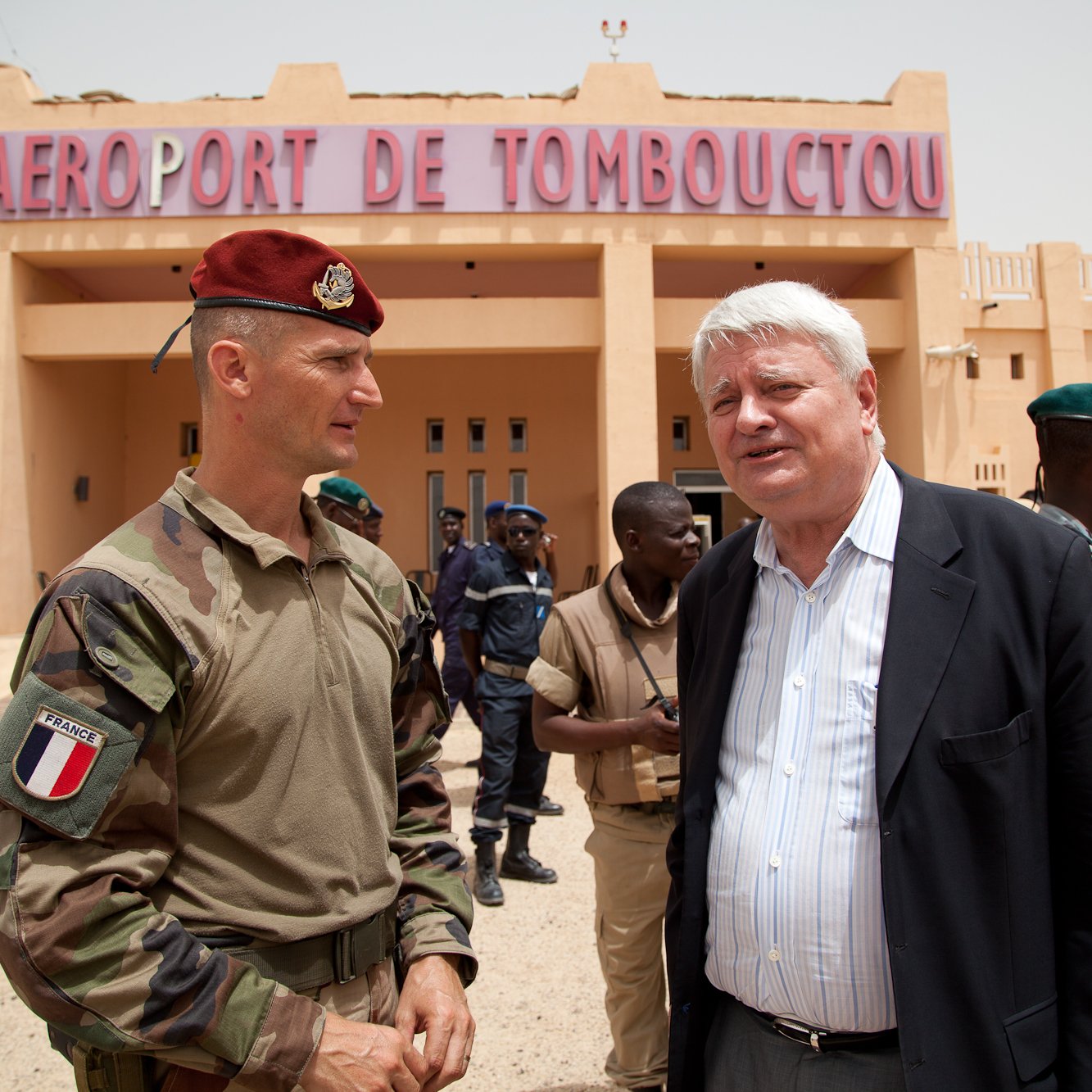 L'image montre une scène à l'aéroport de Tombouctou, avec un bâtiment beige en arrière-plan portant le nom "AÉROPORT DE TOMBOUCTOU" en lettres rouges. Au premier plan, deux hommes sont en conversation. L'un est en uniforme militaire, portant un béret rouge, tandis que l'autre est habillé en costume. Autour d'eux, plusieurs personnes en uniforme, probablement des militaires et des policiers, sont présentes, créant une atmosphère chargée d'activités. Le ciel est clair et le paysage indique une région chaude, avec un sol désertique.
