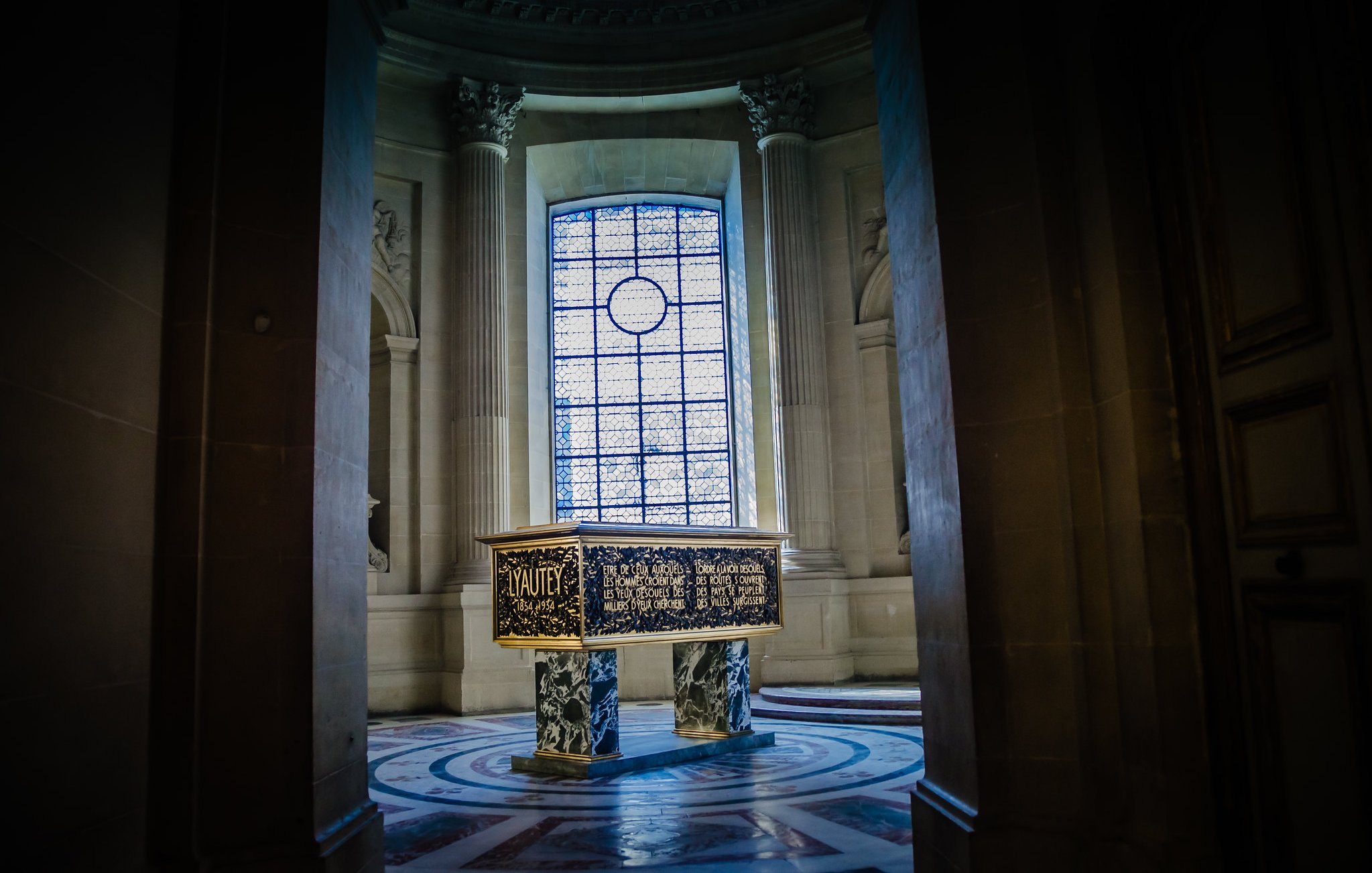 Cette image montre une pièce d'un bâtiment majestueux, probablement un musée ou un palais. Au centre, se trouve un coffre sombre et décoré, surélevé sur un socle circulaire. Les côtés du coffre sont ornés de motifs complexes et d'inscriptions dorées. En arrière-plan, une grande fenêtre ornée de vitraux diffuse une lumière douce qui éclaire la pièce. Les murs sont en pierre grise, enrichis de colonnes majestueuses. L'atmosphère générale est à la fois calme et solennelle, invitant à la contemplation.