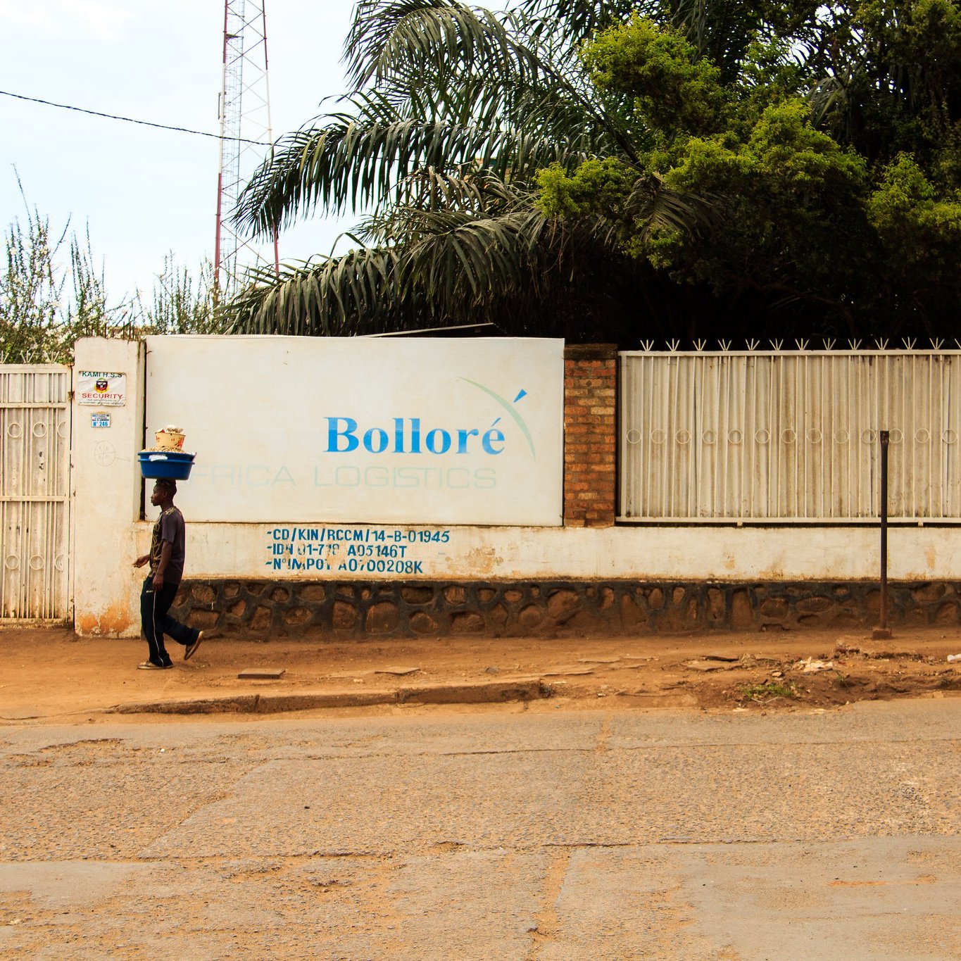 L'image montre un paysage urbain avec une route en terre battue. Sur le côté gauche, une personne marchant lentement porte un fardeau sur sa tête, ce qui donne une impression de travail acharné. À droite, un grand mur blanc affiche le nom "Bolloré" ainsi que des inscriptions et des informations liées à l'entreprise, à côté d'une clôture en métal. En arrière-plan, quelques arbres et une tour de communication sont visibles, ajoutant une touche de verdure à la scène. L'atmosphère semble calme, avec un éclairage naturel qui suggère une journée ensoleillée.