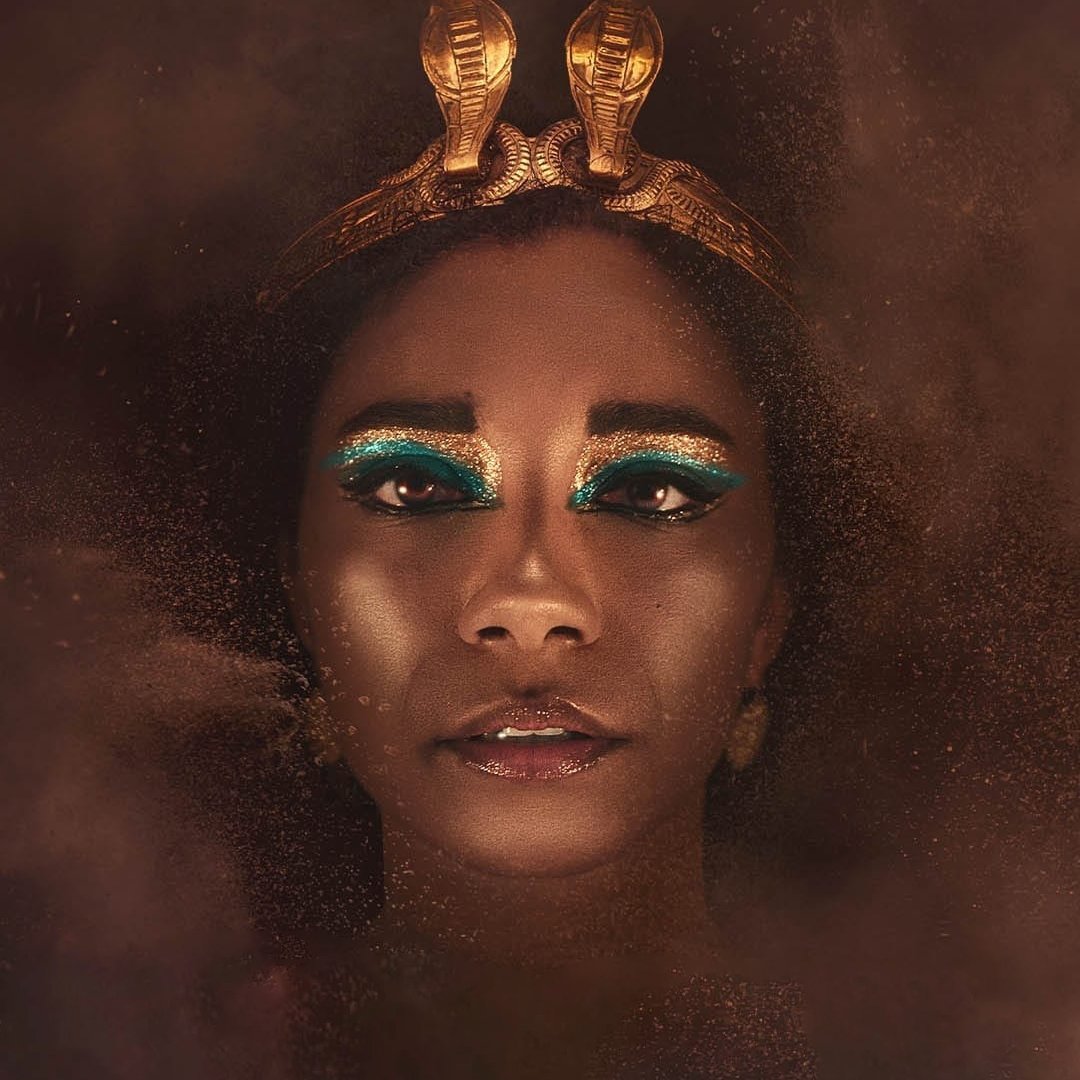 L'image montre un portrait d'une femme avec une peau sombre et un regard intense. Elle porte une couronne dorée ornée de serpents. Son maquillage se distingue par des teintes vibrantes de turquoise autour des yeux, ajoutant un contraste frappant à son visage. La lumière douce crée une ambiance mystérieuse, tandis qu'un brouillard léger autour d'elle donne une impression d'éthéré. L'ensemble dégage une aura de majesté et de puissance.