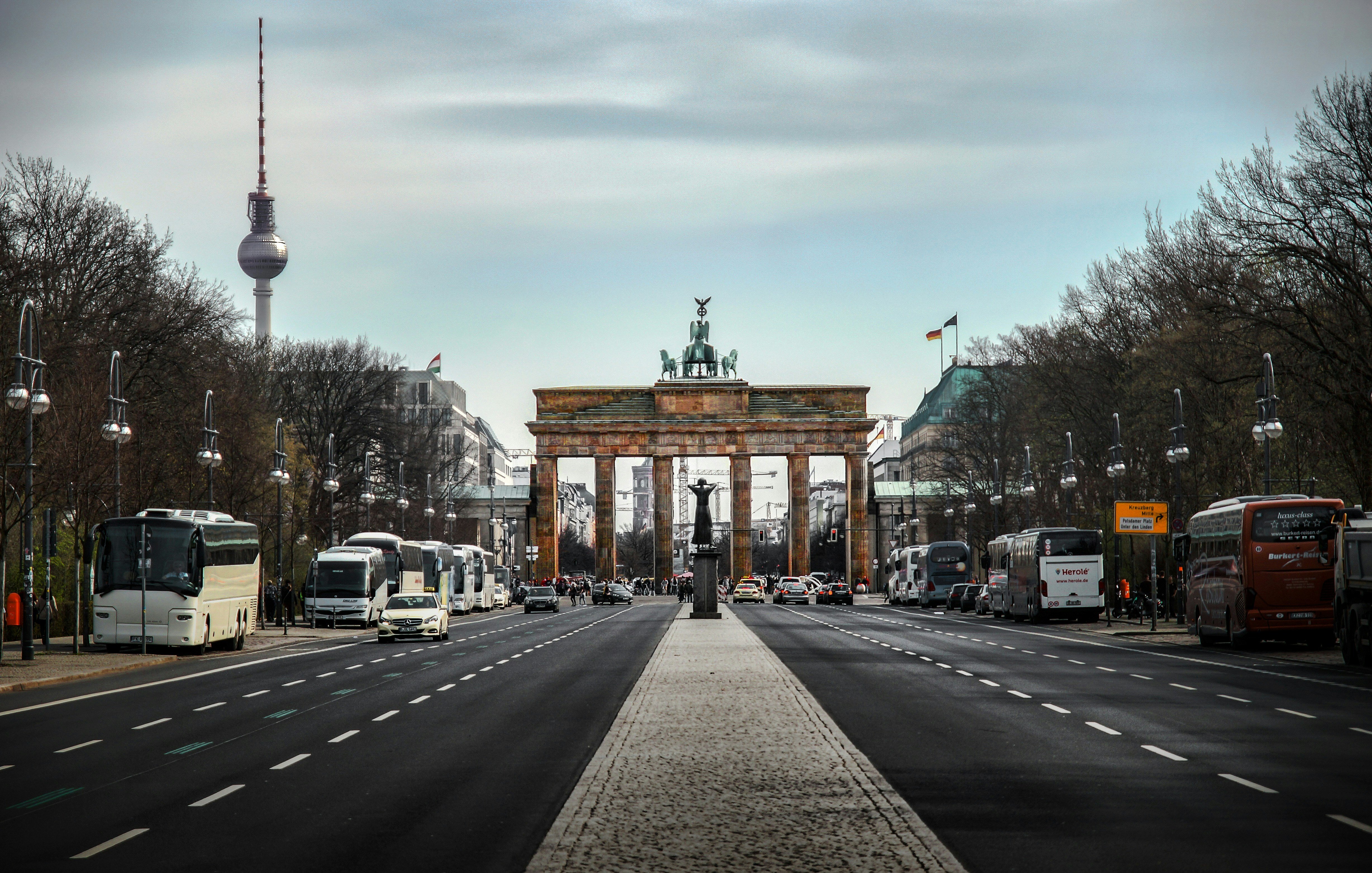 Cette image représente un point de vue d'une grande avenue à Berlin, avec en toile de fond la célèbre porte de Brandebourg. Cette porte emblématique, ornée d'une sculpture représentant un char tiré par des chevaux, est encadrée par des arbres dénudés, typiques des saisons froides. À gauche, on aperçoit la tour de télévision de Berlin, un autre symbole de la ville, se dressant majestueusement dans le ciel. L'avenue est bordée de bus et de véhicules, et la chaussée est pavée de dalles claires, créant un contraste agréable avec le bitume. Le ciel est légèrement nuageux, ajoutant une ambiance calme et réfléchie à cette scène urbaine.