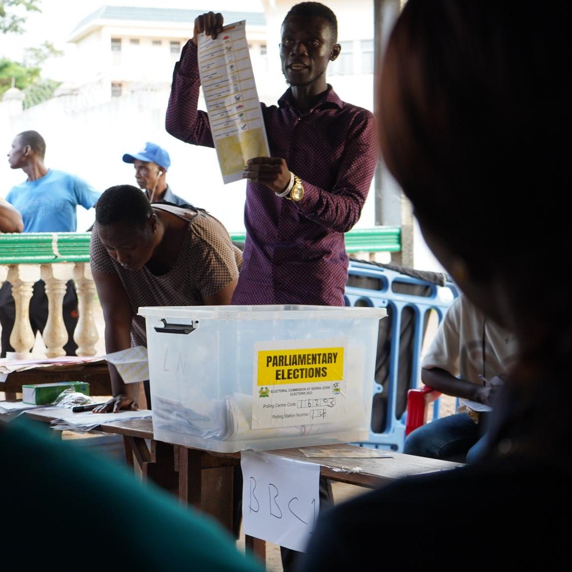 The image depicts a scene from a polling station during a parliamentary election. A man, likely an election official, is holding up a ballot sheet for voters to see. In front of him is a transparent box labeled "PARLIAMENTARY ELECTIONS," suggesting it's used for collecting votes. In the background, several people are gathered, observing the process, while some focus on the official. The setting appears to be outdoors, possibly in a community area, and there's a sense of engagement and participation in the democratic process.