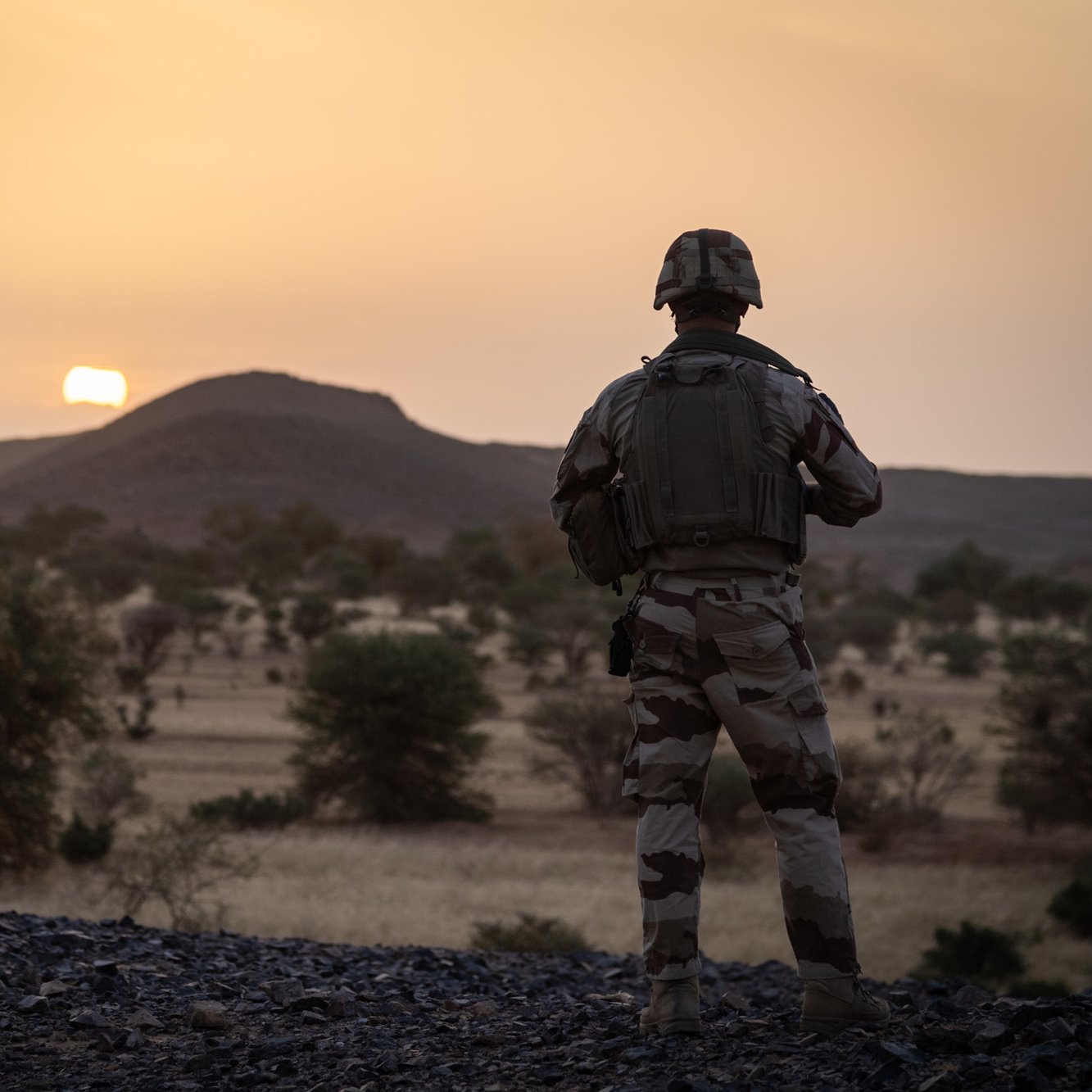 La scène se déroule dans un paysage désertique au coucher du soleil. Au premier plan, un soldat en uniforme camouflage se tient debout, tourné vers l'horizon. Sa silhouette est mise en valeur par la lumière dorée du soleil qui descend lentement derrière une colline arrondie. Le terrain est rocailleux et parsemé de petites végétations et arbres, typiques des paysages arides. L'ambiance est calme, presque contemplative, ajoutant une dimension de réflexion à la présence du soldat, probablement en mission. Les couleurs chaudes du ciel se mélangent à la beauté naturelle du paysage, créant une atmosphère à la fois sereine et intense.