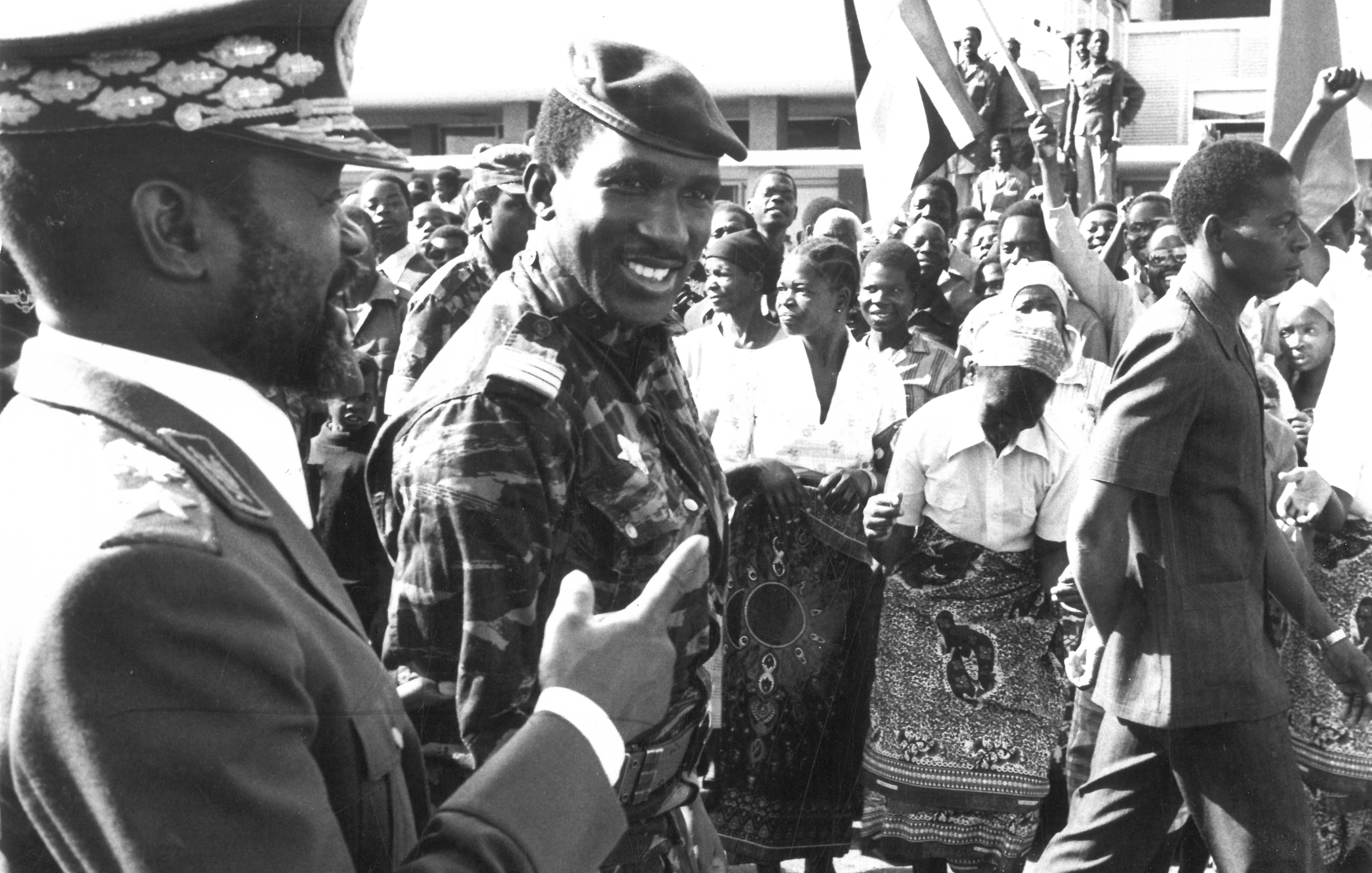 The image depicts a historical scene likely from a public event or rally. In the foreground, two men are engaging in conversation; one is dressed in a military uniform, while the other wears a military-style jacket with insignia. They both seem to be smiling and interacting positively. Behind them, a crowd of people of various ages is visible, with many holding flags and displaying enthusiastic gestures. The atmosphere appears lively, suggesting a moment of celebration or political significance. The image is in black and white, adding to its historical context.