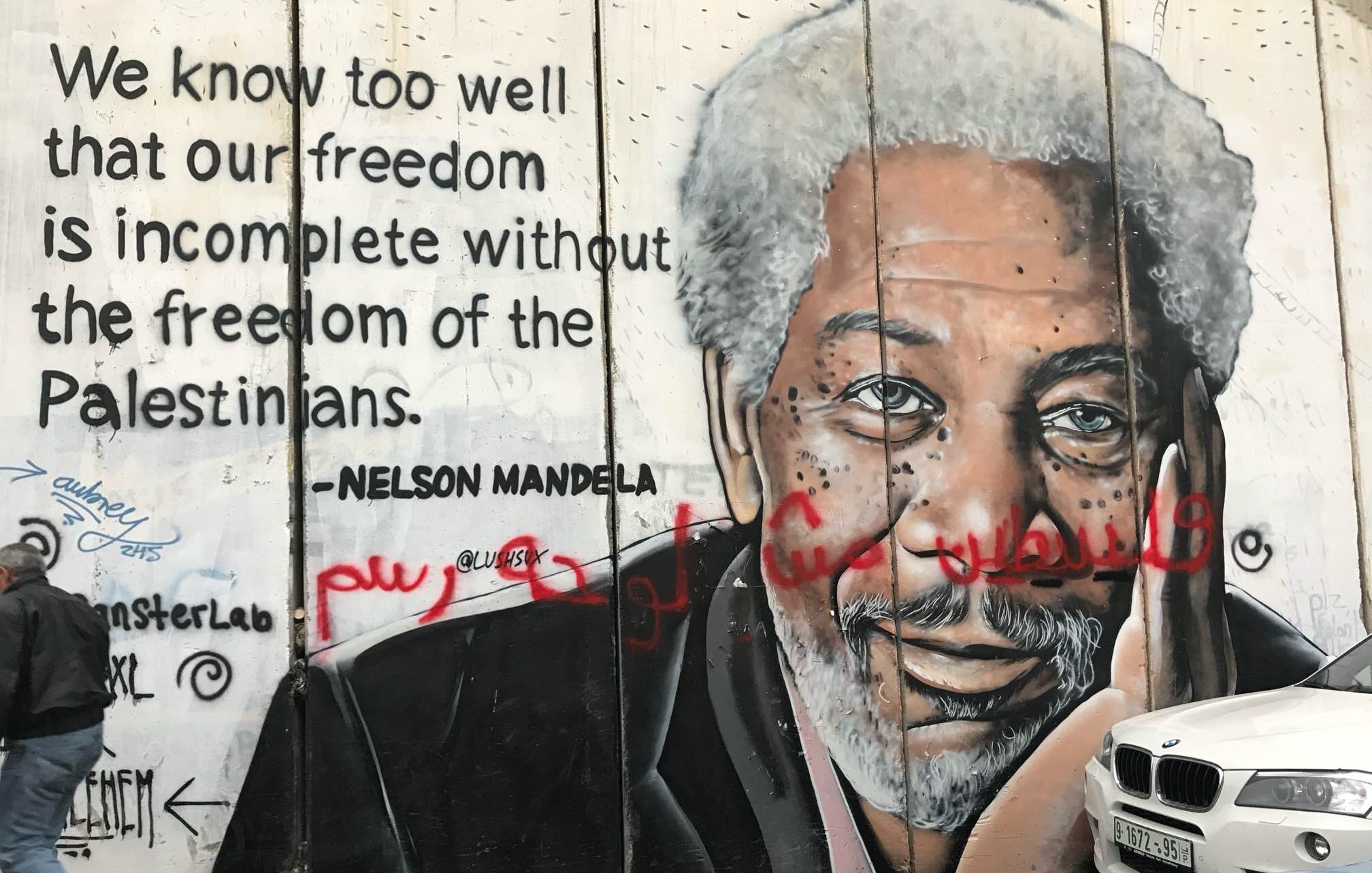 L'image représente un mur sur lequel est peint un mural poignant. À gauche, on voit un portrait détaillé d'un homme âgé avec des cheveux grisonnants et une expression réfléchie. À droite du portrait, une citation attribuée à Nelson Mandela évoque l'importance de la liberté des Palestiniens pour garantir notre propre liberté. Le texte est en anglais, avec un style graphique qui attire l'attention. En bas, quelques graffitis ajoutent une touche de couleur et de dynamisme à l'ensemble. L'ambiance est à la fois engageante et réfléchie, signifiant une connexion profonde entre les luttes pour la liberté.