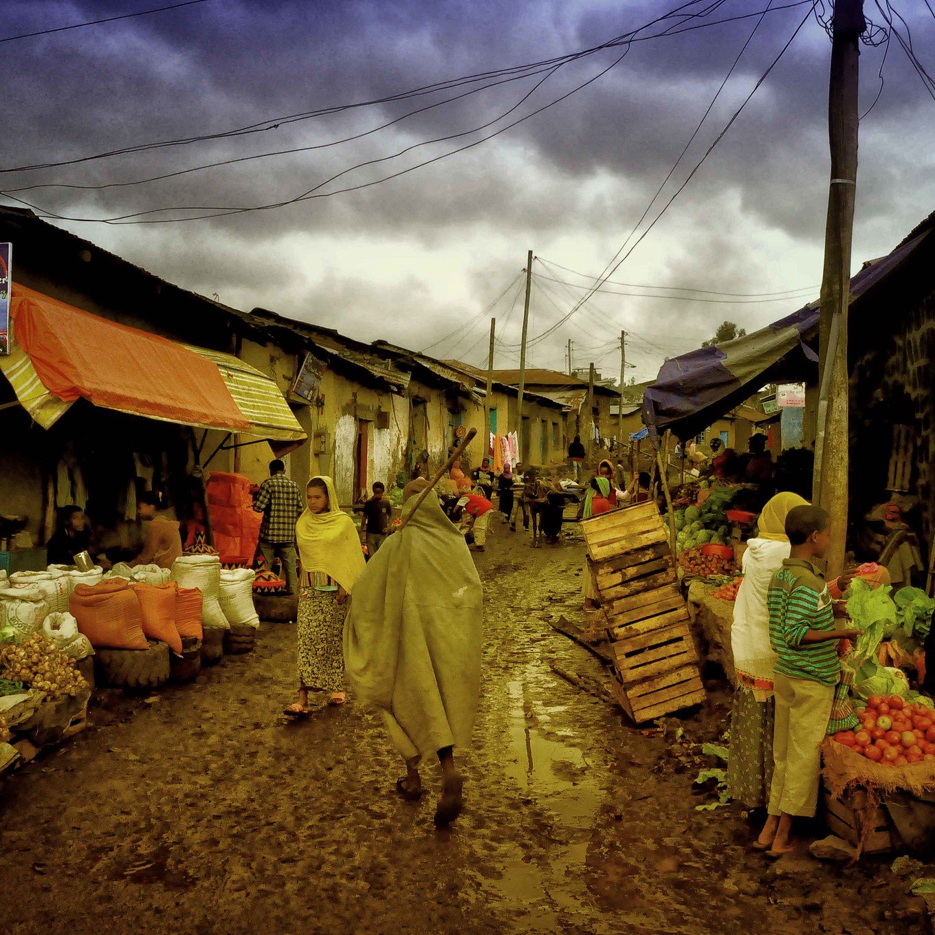 Cette image dépeint une scène de marché dans une rue étroite et animée. Le sol est boueux, probablement à cause de la pluie récente. Des étals colorés, couverts de bâches orange et bleues, sont remplis de fruits et légumes frais. Des vendeurs, portant des vêtements traditionnels et des capuchons pour se protéger de l'humidité, interagissent avec les clients. On peut ressentir l'effervescence du marché, avec des gens qui marchent, négocient et échangent des sourires. Au fond, le ciel est nuageux, ajoutant une atmosphère grise à cette scène vivante. Ce mélange de sons, d'odeurs et d'activités crée une ambiance dynamique et chaleureuse, typique des marchés locaux.