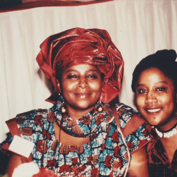 L'image montre deux femmes souriantes posant côte à côte. La femme à gauche porte un vêtement traditionnel aux motifs colorés, accompagné d'un foulard assorti. Sa expression est douce et chaleureuse. À sa droite, la seconde femme affiche également un grand sourire ; elle porte un haut à motifs et des accessoires qui ajoutent une touche élégante à son apparence. L'environnement semble être festif, avec une nappe blanche en arrière-plan, laissant planer une atmosphère accueillante et joyeuse.