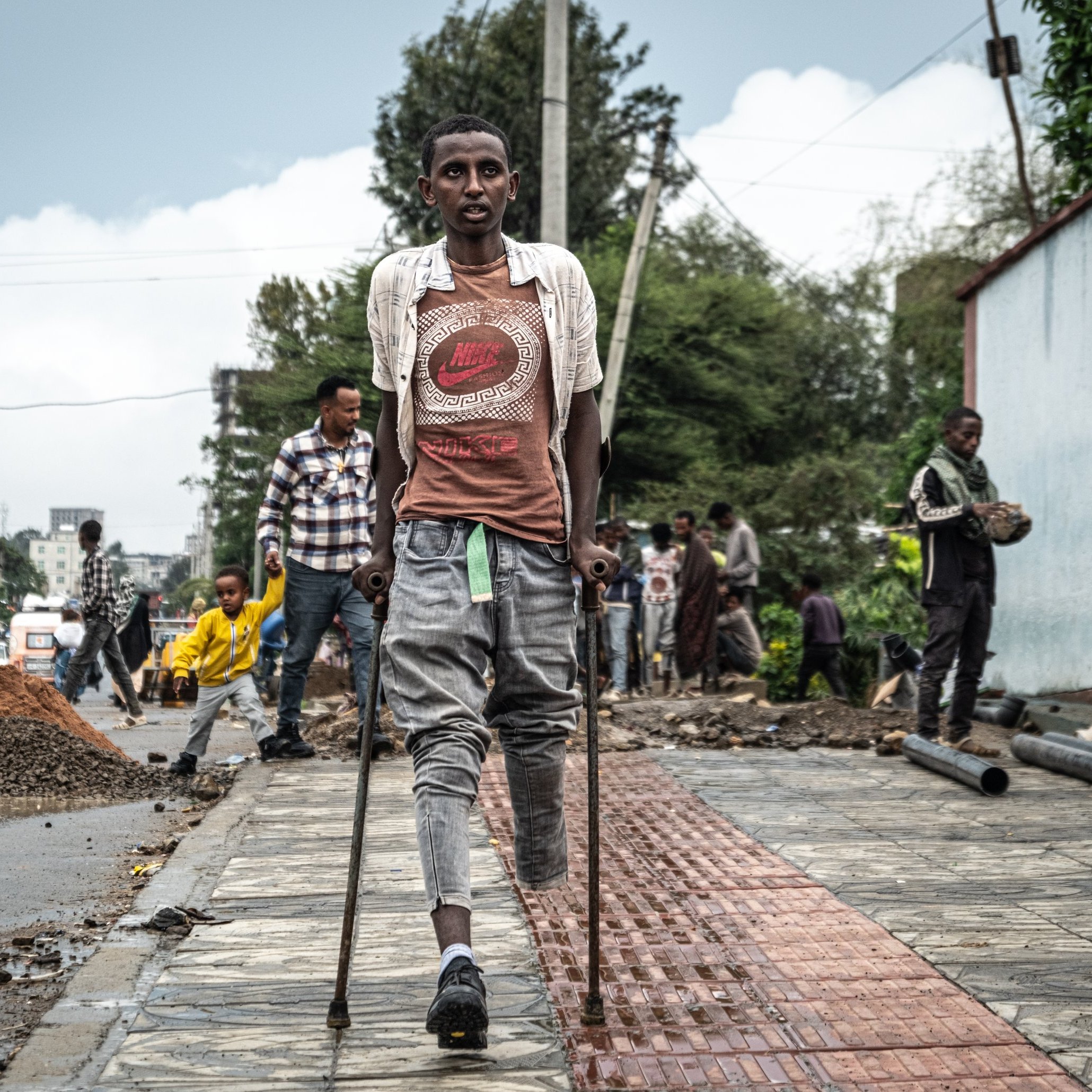 L'image montre une scène urbaine animée, où plusieurs personnes sont visibles dans un environnement de construction. Au premier plan, un jeune homme marche avec des béquilles, portant un t-shirt et un short. Il semble concentré et déterminé. En arrière-plan, on aperçoit d'autres travailleurs s'affairant dans la rue, entourés de matériaux de construction comme du gravier et des tuyaux. Le ciel est nuageux, suggérant une atmosphère légèrement grise. À droite, un mur peint est visible, ajoutant une touche de couleur au paysage de chantier.