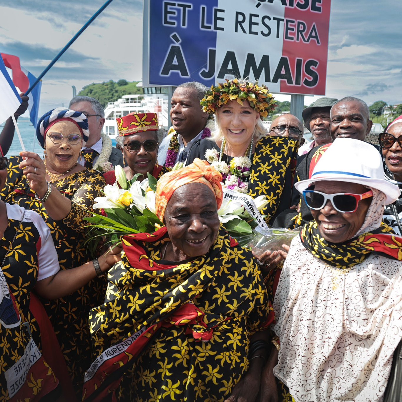 L'image représente une scène festive, probablement lors d'une célébration culturelle ou d'un événement politique. Au centre, plusieurs femmes et hommes portent des vêtements colorés, souvent ornés de motifs floraux jaunes sur fond noir. Certains participants tiennent des bouquets de fleurs tandis que d'autres brandissent des drapeaux français. Au fond, un grand panneau affiche un message qui semble souligner un attachement fort à la France, avec les mots "ET LE RESTERA À JAMAIS". L'atmosphère est joyeuse, avec des sourires et des expressions de célébration visible sur les visages des participants. Le ciel en arrière-plan est partiellement nuageux, ajoutant à l'ambiance vibrante de l'événement.