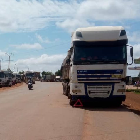 L'image montre une route large et droite, où une file de camions est stationnée sur le côté. Les camions, de différentes tailles et couleurs, sont alignés de manière ordonnée. Le ciel est clair avec quelques nuages blancs qui flottent. On peut apercevoir des poteaux électriques le long de la route, et au loin, il y a des bâtiments, suggérant un environnement urbain en arrière-plan. À proximité, un motocycliste passe, ajoutant un peu de dynamisme à la scène calme. L'ambiance générale dégage une impression de circulation routière et d'activités commerciales.