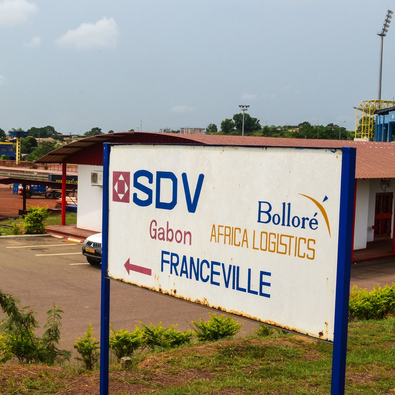 L'image montre un panneau indicatif dans une zone logistique. Ce panneau, situé devant un bâtiment, indique la direction vers Franceville, une ville du Gabon. On peut apercevoir le logo de SDV, ainsi que celui de Bolloré Africa Logistics. Le paysage environnant est assez verdoyant, avec plusieurs arbres et buissons. Au loin, on distingue une infrastructure de transport, possiblement liée à la logistique. L'atmosphère est calme, reflétant un lieu dédié aux activités commerciales et de logistique.