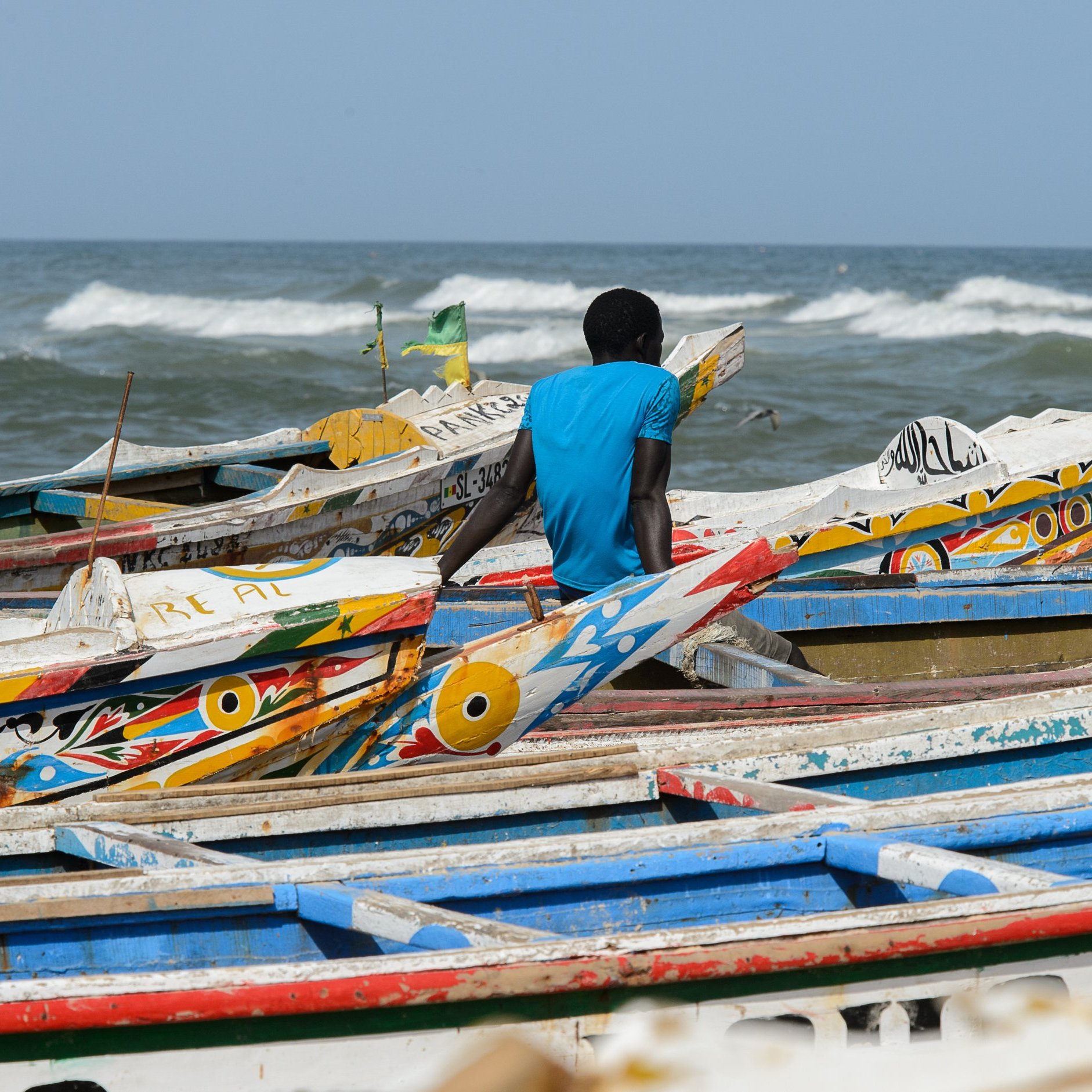 L'image montre une plage animée au bord de la mer. Des bateaux colorés se chevauchent sur le sable, avec des motifs vifs et des teintes variées, tels que le bleu, le rouge et le jaune. Un homme, vêtu d'un t-shirt bleu, est assis au bord des bateaux, tourné vers l'océan. Les vagues s'écrasent doucement sur le rivage, créant un bruit apaisant. Dans l'air, on peut sentir une brise marine fraîche et éventuellement entendre le cri des oiseaux qui volent autour. L'ambiance dégage une sensation de tranquillité tout en étant dynamique grâce à la présence des bateaux et de l'homme observant l'horizon.