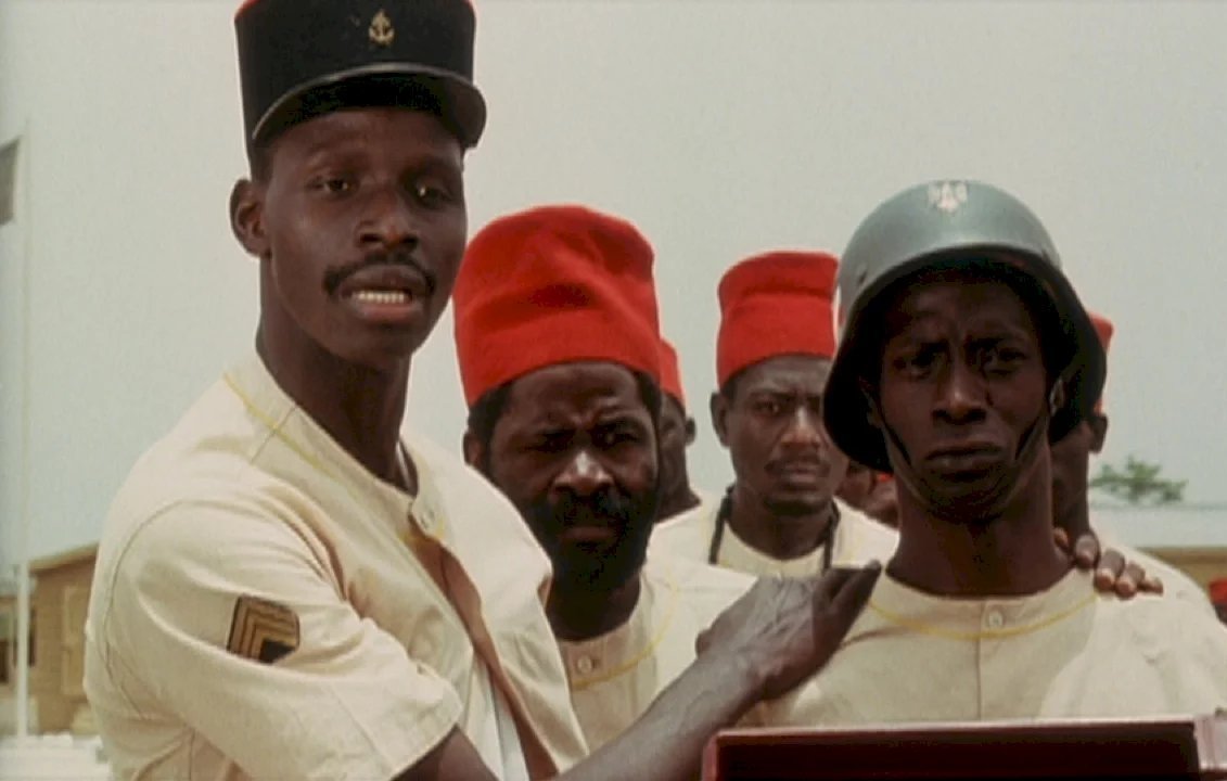 L'image montre plusieurs hommes, en uniformes, rassemblés dans un environnement clair. Au premier plan, un homme se tient à gauche, portant un chapeau noir avec un insigne et une tenue beige. Il regarde vers la droite avec une expression sérieuse. À côté de lui, un autre homme, portant un casque militaire et un uniforme beige, arbore un air grave. Derrière eux, plusieurs autres hommes regardent également dans la même direction. Tous semblent engagés dans une discussion ou une situation importante. Les couleurs dominantes de l'image sont le beige des uniformes et le rouge vif des chapeaux de certains des hommes.