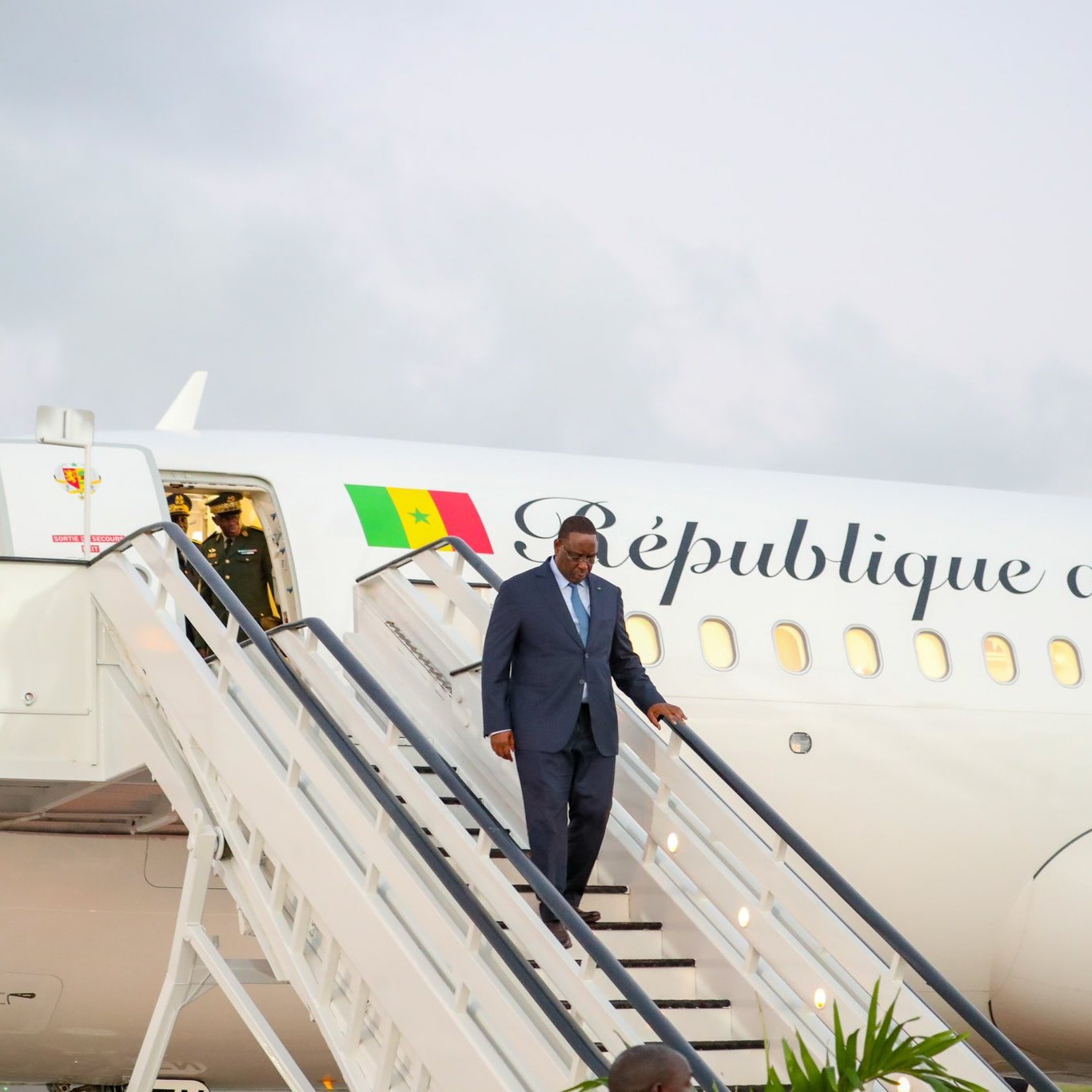 L'image montre un homme en costume sombre descendant d'un avion. L'avion, aux couleurs blanches et avec l'inscription "République du Sénégal" sur le fuselage, est garé sur une piste. À l'arrière-plan, le ciel est légèrement nuageux. Le personnage semble marcher avec assurance, et des escaliers mènent directement à la porte de l'avion. Au sol, on peut voir quelques plantes, suggérant un environnement accueillant.