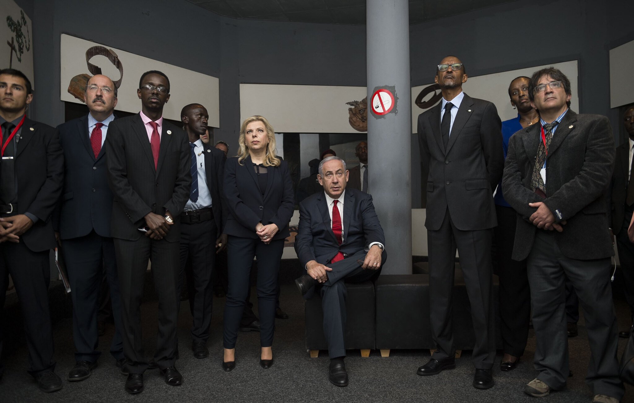 L'image montre un groupe de personnes rassemblées dans une salle. Au centre, un homme est assis sur un canapé, vêtu d'un costume, tandis que les autres se tiennent debout autour de lui. Ils portent tous des costumes et ont des expressions sérieuses, suggérant une atmosphère formelle. L'environnement semble être une exposition ou un espace culturel, avec des œuvres d'art ou des objets exposés sur les murs. L'éclairage est doux, créant une ambiance réfléchie et attentive.