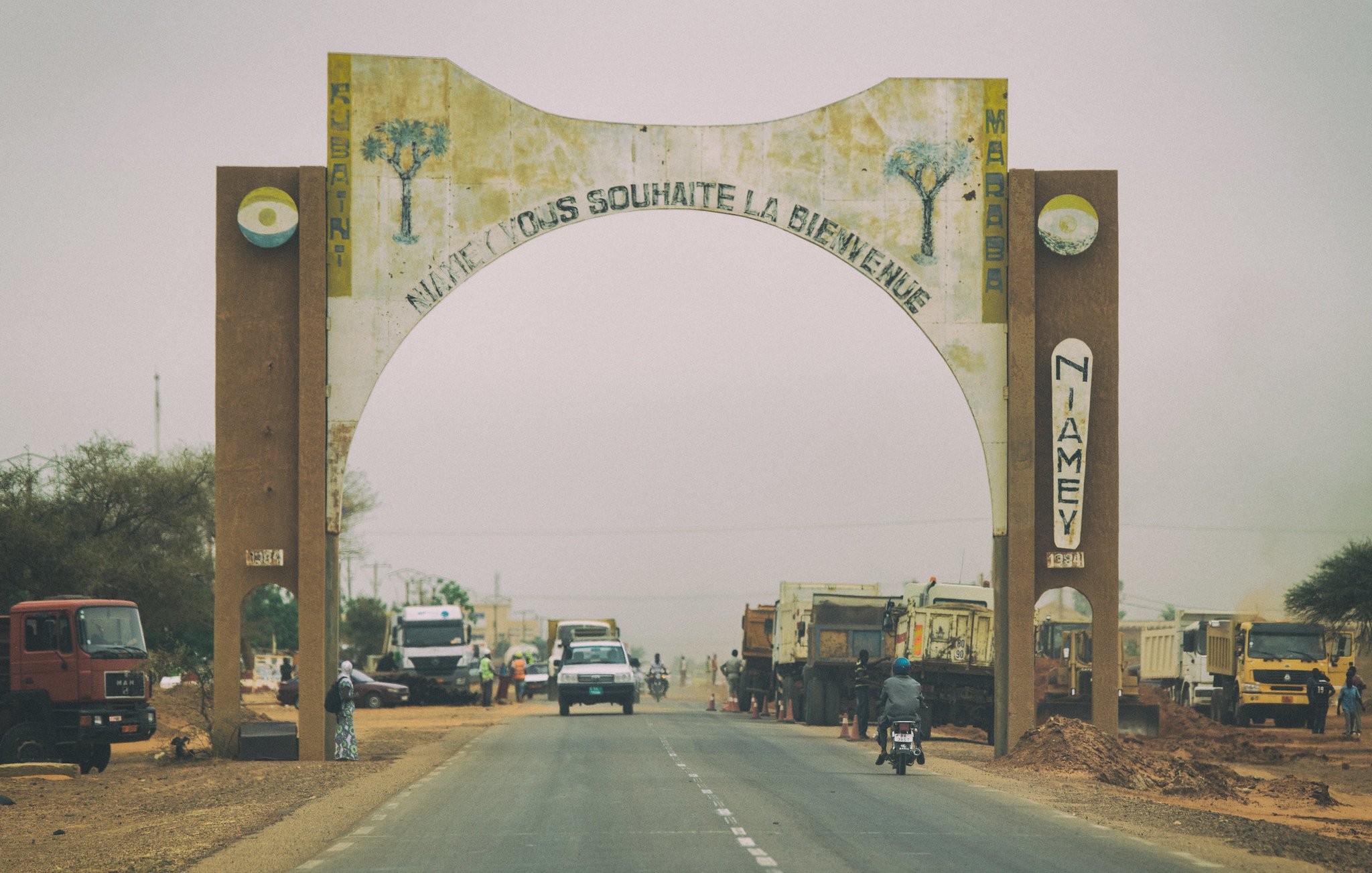L'image montre une route qui traverse une grande arche d'entrée, symbolisant l'arrivée à Niamey, la capitale du Niger. L'arche est décorée avec des motifs végétaux et des inscriptions en français qui souhaitent la bienvenue. De chaque côté de la route, on peut voir des camions et des véhicules qui circulent, ainsi que des arbres et de la terre poussiéreuse. L'atmosphère est légèrement brumeuse, accentuant le caractère désertique de la région. L'ensemble donne une impression dynamique de la vie et du commerce à l'entrée de la ville.