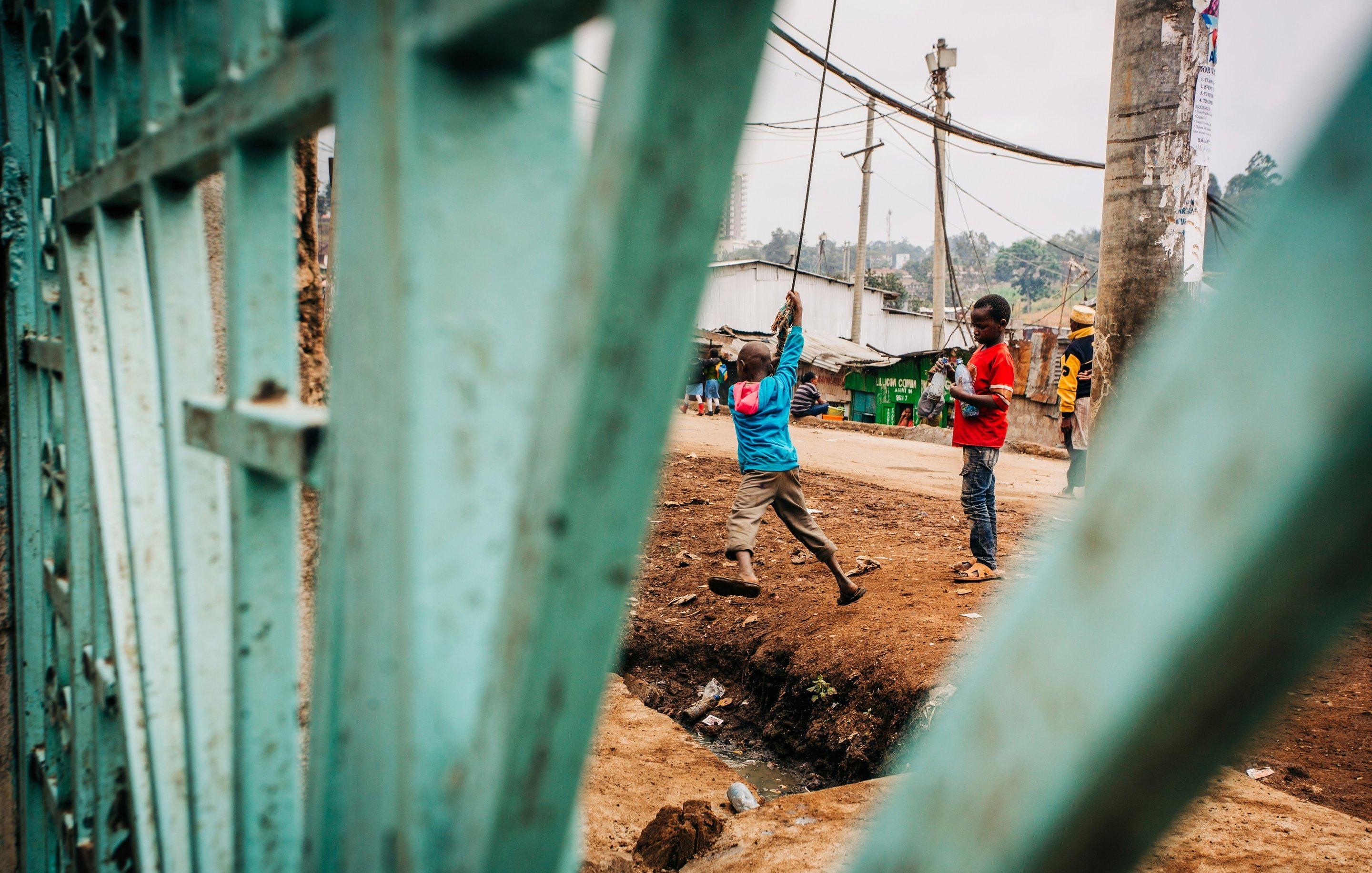 L'image présente une scène de la vie quotidienne dans un environnement urbain. Au premier plan, on aperçoit des barres d'une grille verte, qui encadrent la vue. À l'intérieur, deux enfants jouent près d'un chemin. L'un porte une chemise bleue et tient un objet au-dessus de sa tête, semblant s'amuser. L'autre enfant, aux vêtements rouges, observe ou se prépare à rejoindre le jeu. En arrière-plan, il y a des maisons simples, des poteaux électriques et une ambiance vibrante, typique d'un quartier animé. On sent une atmosphère de camaraderie et d'énergie enfantine. Le terrain est boueux et légèrement en pente.