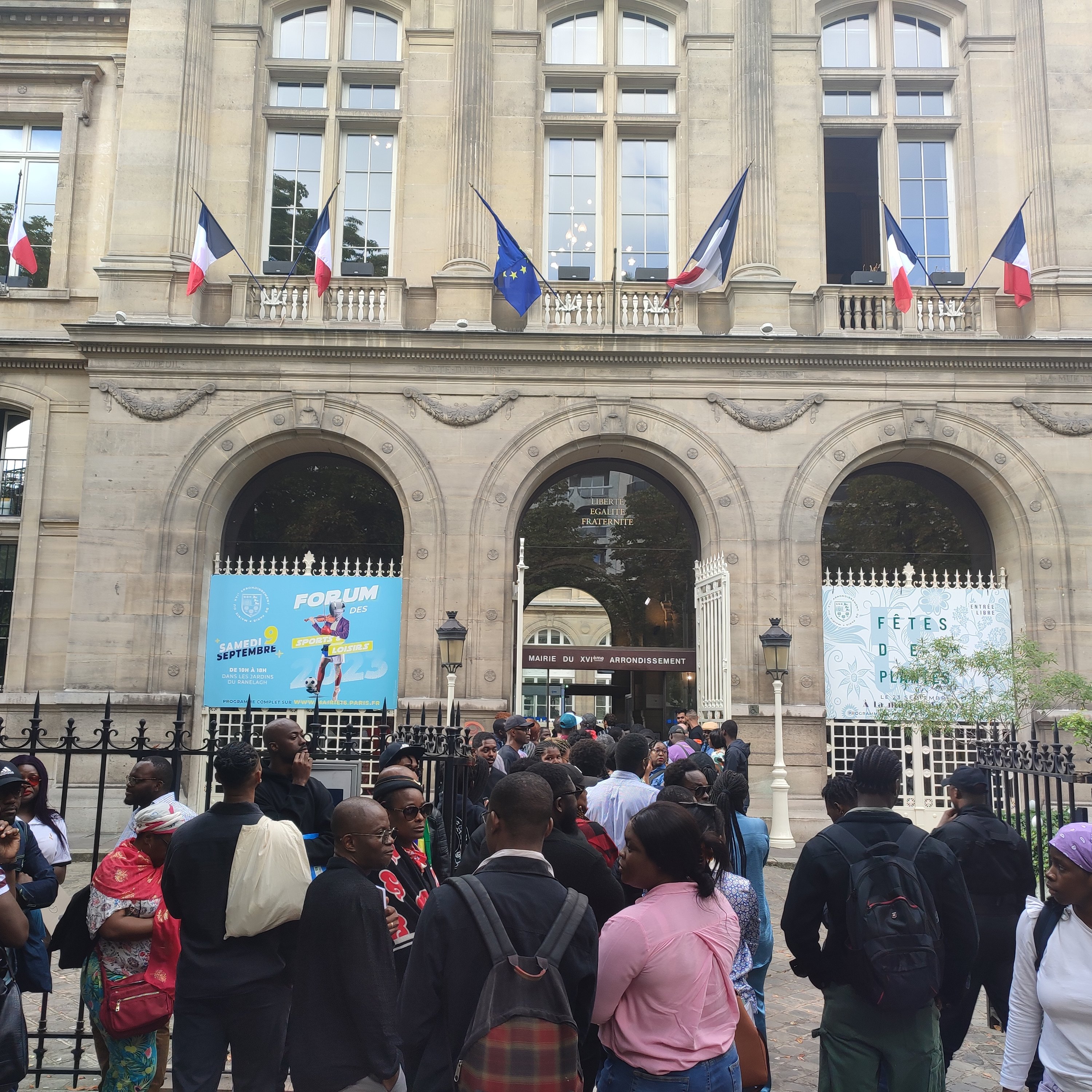 L'image montre un bâtiment imposant avec une architecture classique, visible grâce à ses grandes fenêtres et ses arches. Devant ce bâtiment, un groupe de personnes se rassemble, créant une atmosphère animée. Des drapeaux français flottent au-dessus d'eux, témoignant de l'importance de ce lieu. Les visiteurs semblent variés en termes d'âge et de style vestimentaire, ce qui reflète une diversité culturelle. À l'arrière-plan, des affiches colorées annoncent un événement, ajoutant une touche festive à la scène. L'ensemble dégage une ambiance d'attente et d'excitation.