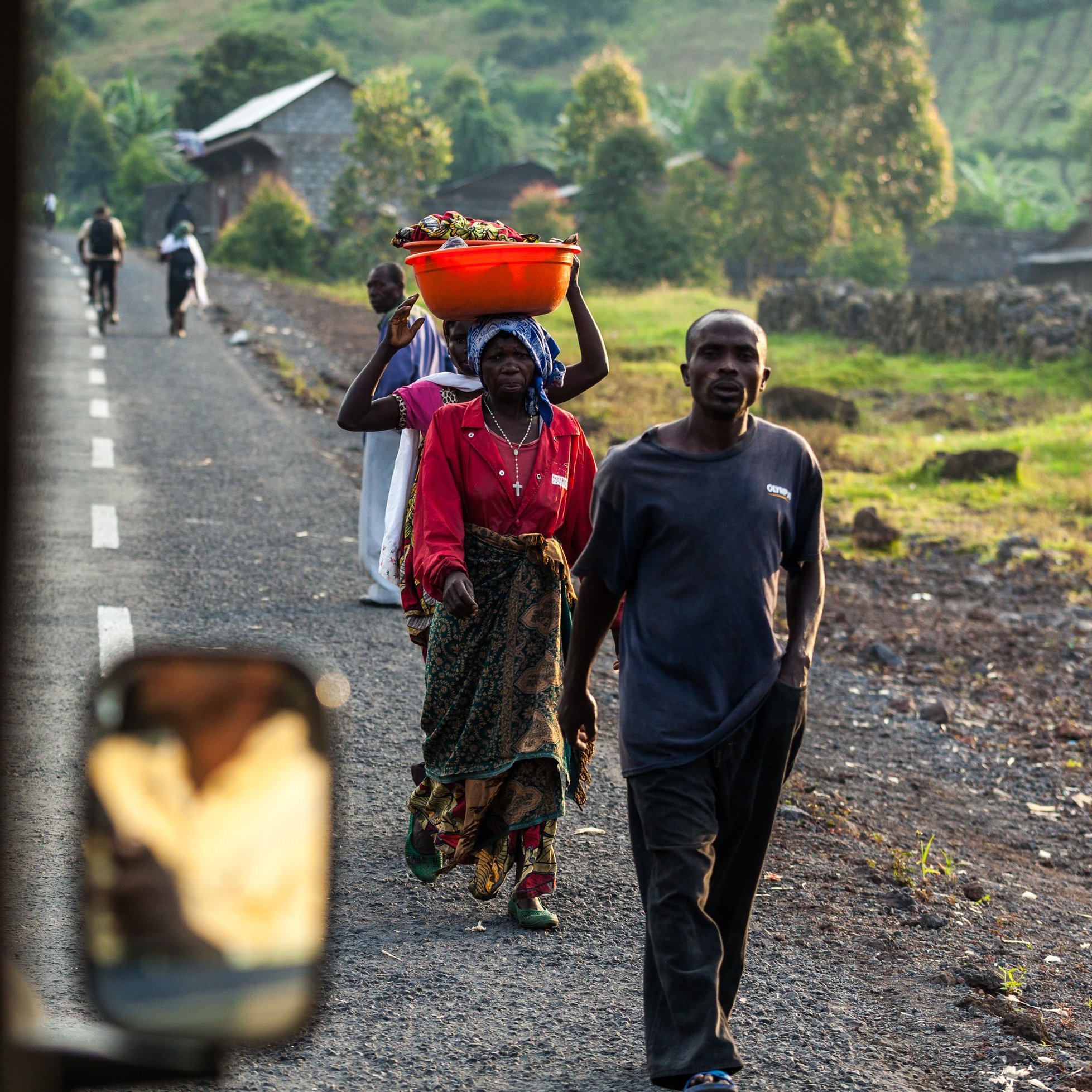 L'image montre une scène de vie quotidienne dans une région rurale. Sur une route en terre, plusieurs personnes marchent, chacune portant des charges sur leurs épaules ou dans les bras. Au premier plan, une femme est vue de dos, portant un grand panier orange rempli de fruits ou de légumes sur sa tête. Elle est vêtue d'une robe colorée. À côté d'elle, un homme porte une veste sombre et marche avec assurance. En arrière-plan, on peut apercevoir d'autres personnes se déplaçant sur la route, ainsi que des habitations et des collines verdoyantes qui donnent une impression de paysage naturel. La lumière du soleil éclaire la scène, créant une atmosphère chaleureuse et vivante.