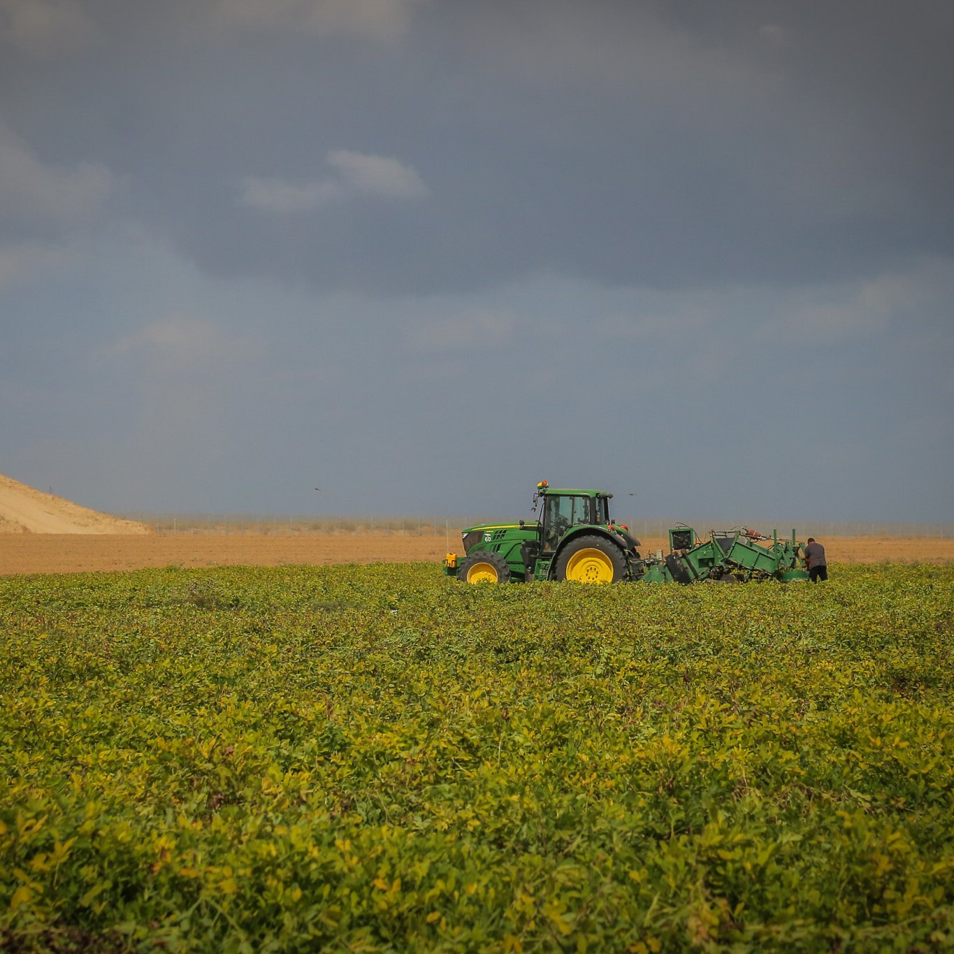 L'image présente un vaste champ de culture où l'on peut voir un tracteur vert, typique des fermes. Le tracteur est engagé dans une activité de labour, préparant le sol pour la plantation. En arrière-plan, on aperçoit une colline de terre argileuse, surmontée d'un véhicule. Le ciel est nuageux, créant une ambiance contrastée avec des teintes de bleu et de gris qui donnent une impression de calme. Les lignes horizontales des cultures et de la colline ajoutent une profondeur visuelle au paysage rural.