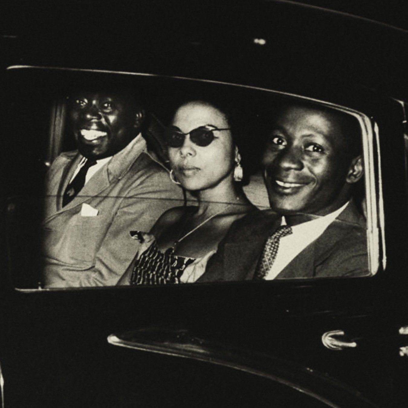 L'image présente trois personnes assises à l'intérieur d'une voiture sombre. L'ambiance semble élégante et vintage. À gauche, un homme souriant, au teint foncé, porte un costume clair. Au centre, une femme avec des lunettes de soleil stylées, habillée d'une robe à motifs, dégage une aura de sophistication. À droite, un autre homme, également souriant, avec un teint légèrement plus clair, porte une tenue qui semble chic. Les visages des occupants sont mis en valeur par un éclairage doux, créant une atmosphère chaleureuse et amicale. Le tout suggère un moment de convivialité, peut-être après une sortie ou un événement.