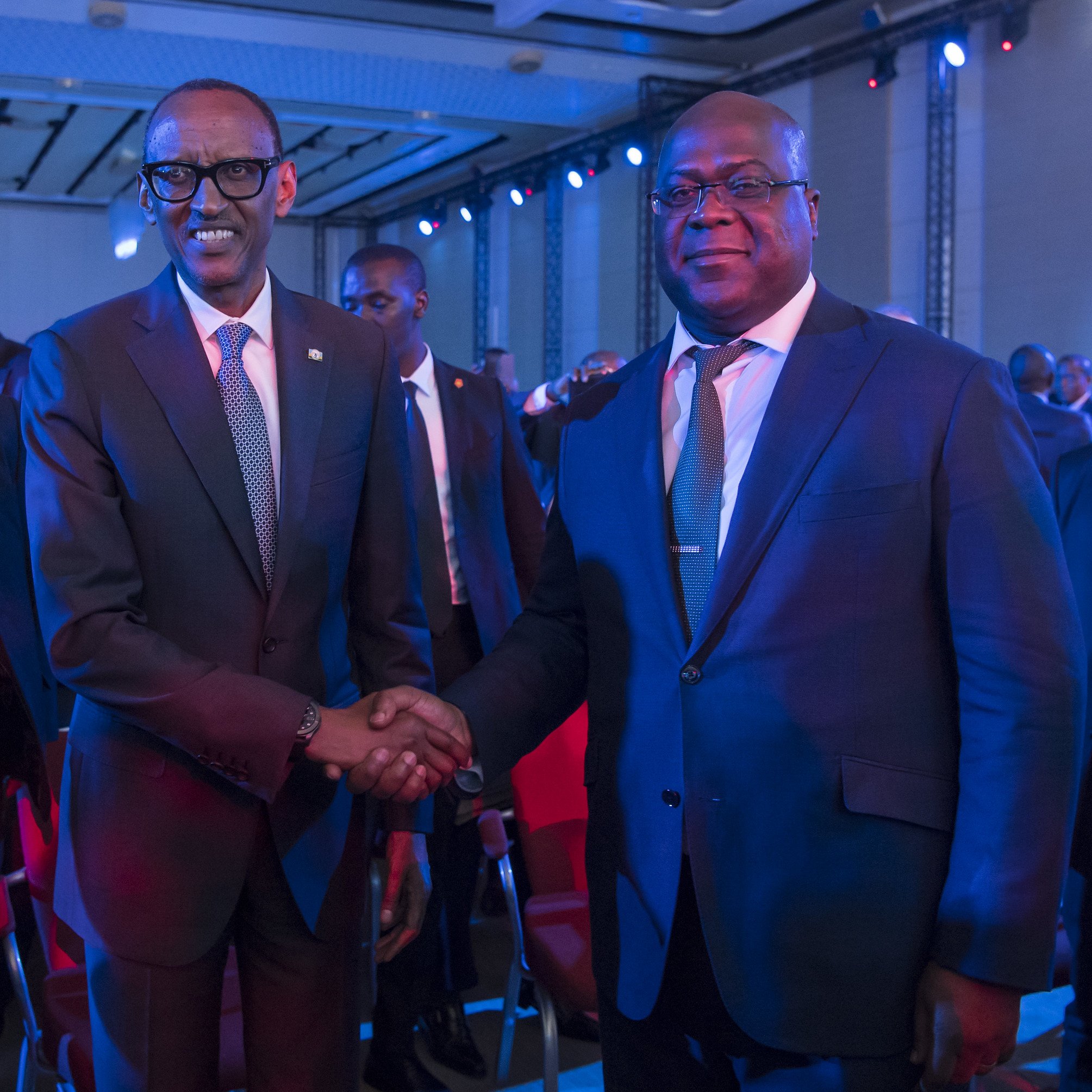 L'image montre deux hommes en costume qui se tiennent debout, se serrant la main dans un contexte de réunion ou d'événement officiel. L'un porte des lunettes et semble d'origine rwandaise, tandis que l'autre est un homme robuste, légèrement plus âgé, probablement d'origine congolaise. Ils affichent des expressions souriantes et amicales, dans une ambiance éclairée par des lumières bleues créant une atmosphère conviviale. En arrière-plan, on peut entrevoir d'autres personnes présentes à l'événement.