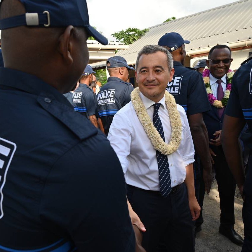Dans cette image, nous voyons un groupe de policiers municipaux, tous vêtus d'un uniforme bleu avec le mot "POLICE" inscrit sur le dos. Au premier plan, un homme souriant échange une poignée de main avec un policier. Cet homme porte une chemise blanche et une cravate, ainsi qu'un collier de fleurs autour du cou, ce qui suggère un événement formel ou culturel. En arrière-plan, on aperçoit d'autres personnes, dont certaines semblent également habillées de manière soignée. La scène se déroule dans un environnement extérieur, avec des bâtiments aux toits en tôle. L'atmosphère semble accueillante et amicale.