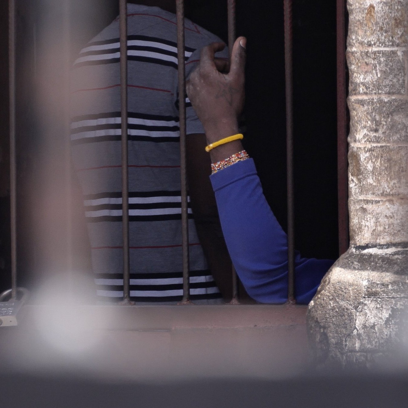 Cette image montre une scène à l'intérieur d'un espace confiné, avec des barreaux visibles au premier plan. On distingue des silhouettes humaines derrière les barreaux. Une main, portant un bracelet, s'élève légèrement, témoignant d'un geste de communication ou de contact. Les murs autour des barreaux semblent anciens et en pierre, ajoutant un élément d'authenticité à l'environnement. L'éclairage suggère que la scène se déroule à l'extérieur, avec une lumière naturelle qui éclaire partiellement l'espace.