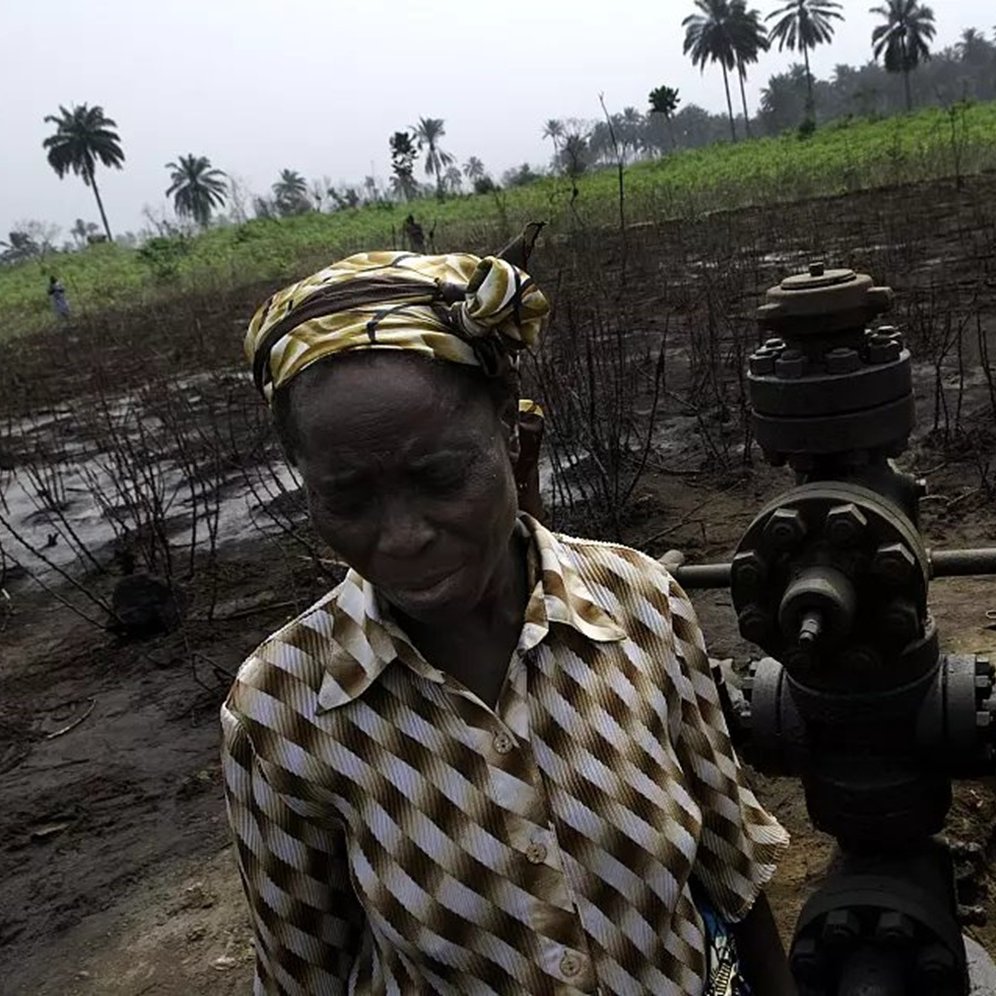 L'image montre un paysage rural où se trouvent des plantations de palmiers en arrière-plan. Au premier plan, une femme d'âge mûr, vêtue d'une blouse rayée et portant un foulard coloré, se tient près d'une infrastructure de forage pétrolier. Son expression est sérieuse et préoccupée. À l'arrière-plan, une autre personne, également en tenue décontractée, semble s'occuper de l'équipement. Le sol est noirci, suggérant une dégradation environnementale. L'atmosphère générale évoque une lutte pour préserver leur lien avec la terre face aux impacts de l'exploitation pétrolière.