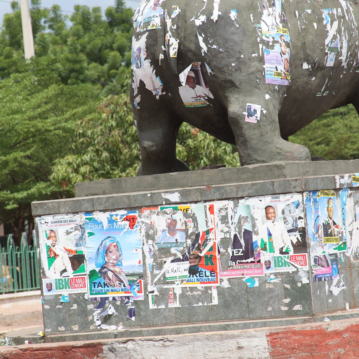 The image depicts a statue, likely an elephant, that is visibly weathered and covered in numerous political posters and flyers. These posters, in various colors, are peeling and overlapping, suggesting a history of political campaigns or events. The statue is situated in an outdoor area, surrounded by greenery and a fence in the background. The overall scene conveys a sense of urban life and the passage of time through the accumulation of posters.