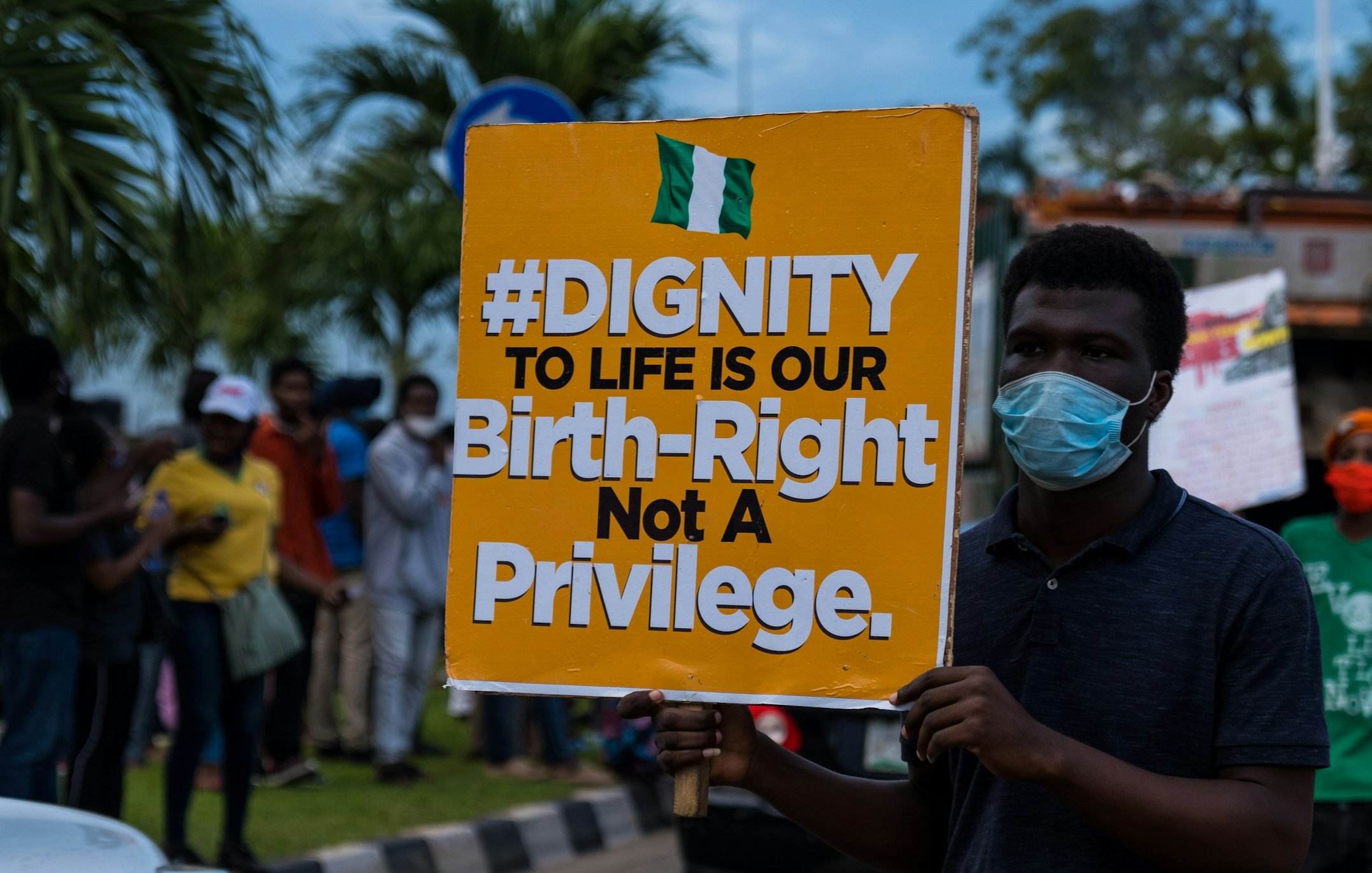 Sur l'image, une manifestation a lieu dans la rue. Un homme se tient au premier plan, portant un masque. Il tient une grande pancarte colorée qui affiche le message suivant : "#DIGNITY TO LIFE IS OUR Birth-Right Not A Privilege" en lettres grandes et noires sur un fond orange. En arrière-plan, on aperçoit un groupe de personnes, certaines portant également des masques, rassemblées autour de lui. L'environnement est urbain, avec des arbres et des véhicules visibles. L'atmosphère est engagée, témoignant d'une lutte pour les droits et la dignité humaine.