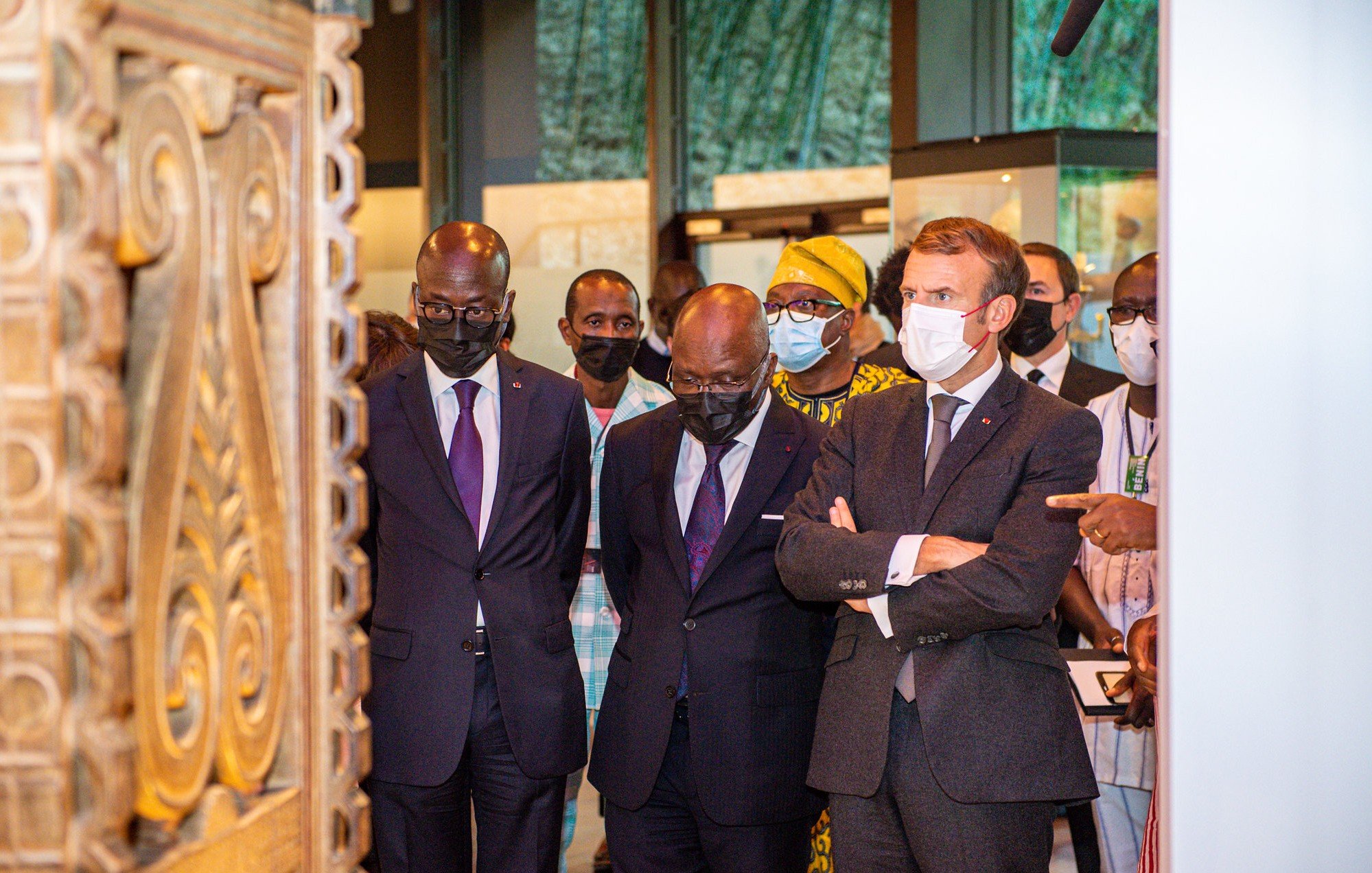 L'image montre un groupe de personnes, dont trois hommes, en train d'observer un objet d'art ou une exposition. Les hommes sont habillés de costumes formels, avec deux en costumes sombres et un en costume gris avec une cravate. Ils affichent des expressions de concentration et d'intérêt. À l'arrière-plan, on perçoit des éléments culturels, probablement des œuvres d'art ou des artifacts, créant une ambiance muséale. Les visiteurs portent tous des masques, ce qui indique qu'ils se trouvent dans un contexte où cela est nécessaire, peut-être en raison de préoccupations sanitaires. Le cadre est bien éclairé, mettant en valeur l'importance de l'exposition.