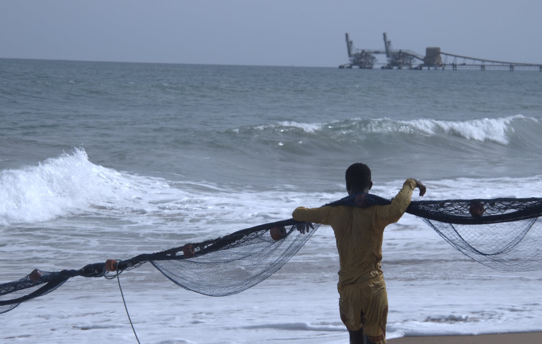 Sur cette image, un homme se trouve sur une plage, dos à nous. Il porte un maillot de bain jaune et tire un grand filet de pêche, qui s'étend devant lui. Les vagues de l'océan s'écrasent doucement sur le sable, produisant un bruit apaisant. À l'arrière-plan, on aperçoit une jetée où se trouvent des structures industrielles, ajoutant une touche de la vie maritime. Le ciel est clair et lumineux, créant une ambiance de tranquillité et de travail en harmonie avec la mer.