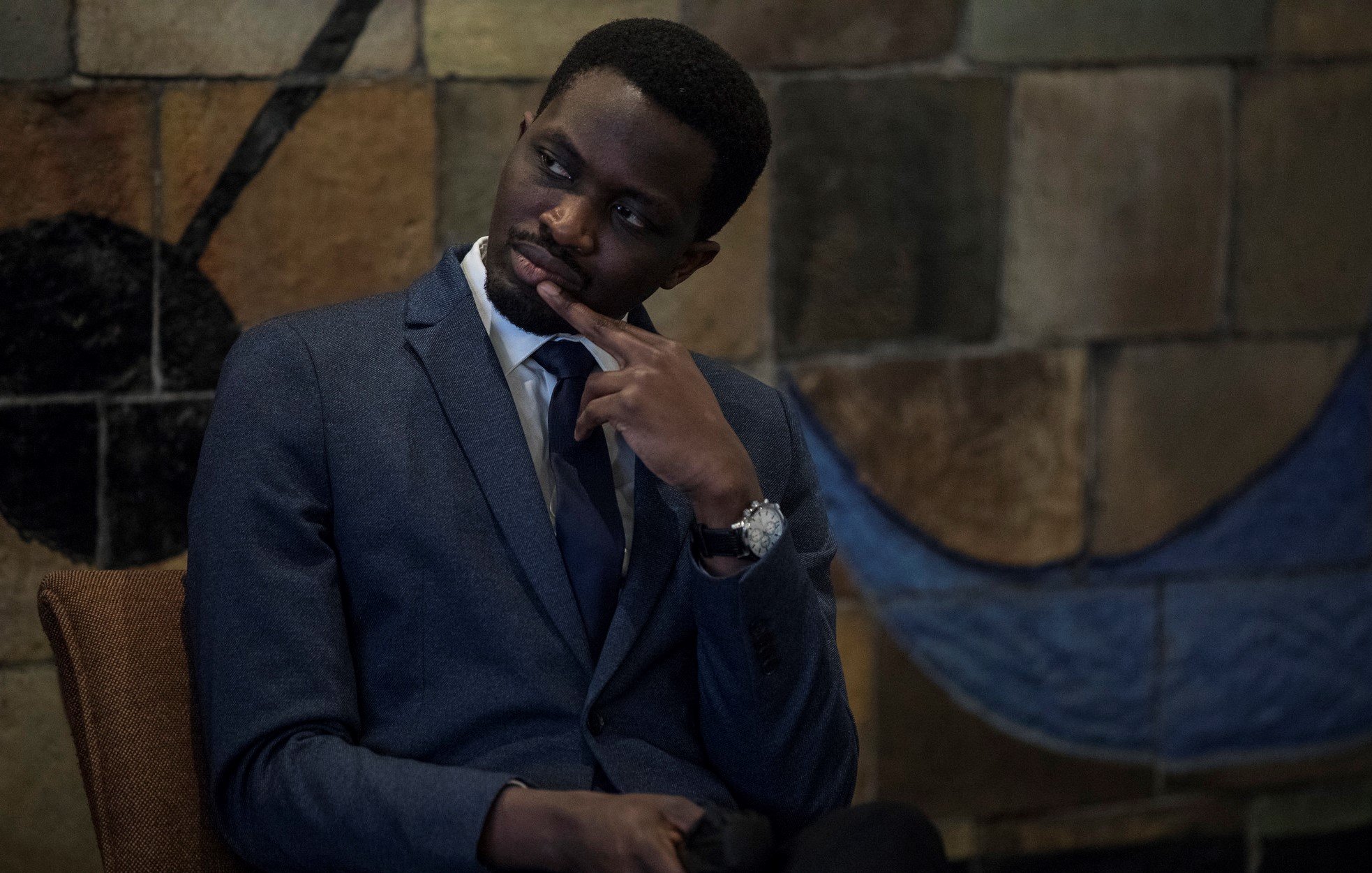 L'image montre un homme assis dans un environnement intérieur. Il porte un costume sombre avec une chemise claire et une cravate, ce qui dénote un certain formalisme. Son expression est pensive, avec un doigt sur le menton, suggérant qu'il réfléchit à quelque chose de sérieux. En arrière-plan, on aperçoit un mur aux motifs colorés, avec des formes géométriques qui ajoutent une touche artistique à la scène. L'ambiance est calme et réfléchie.