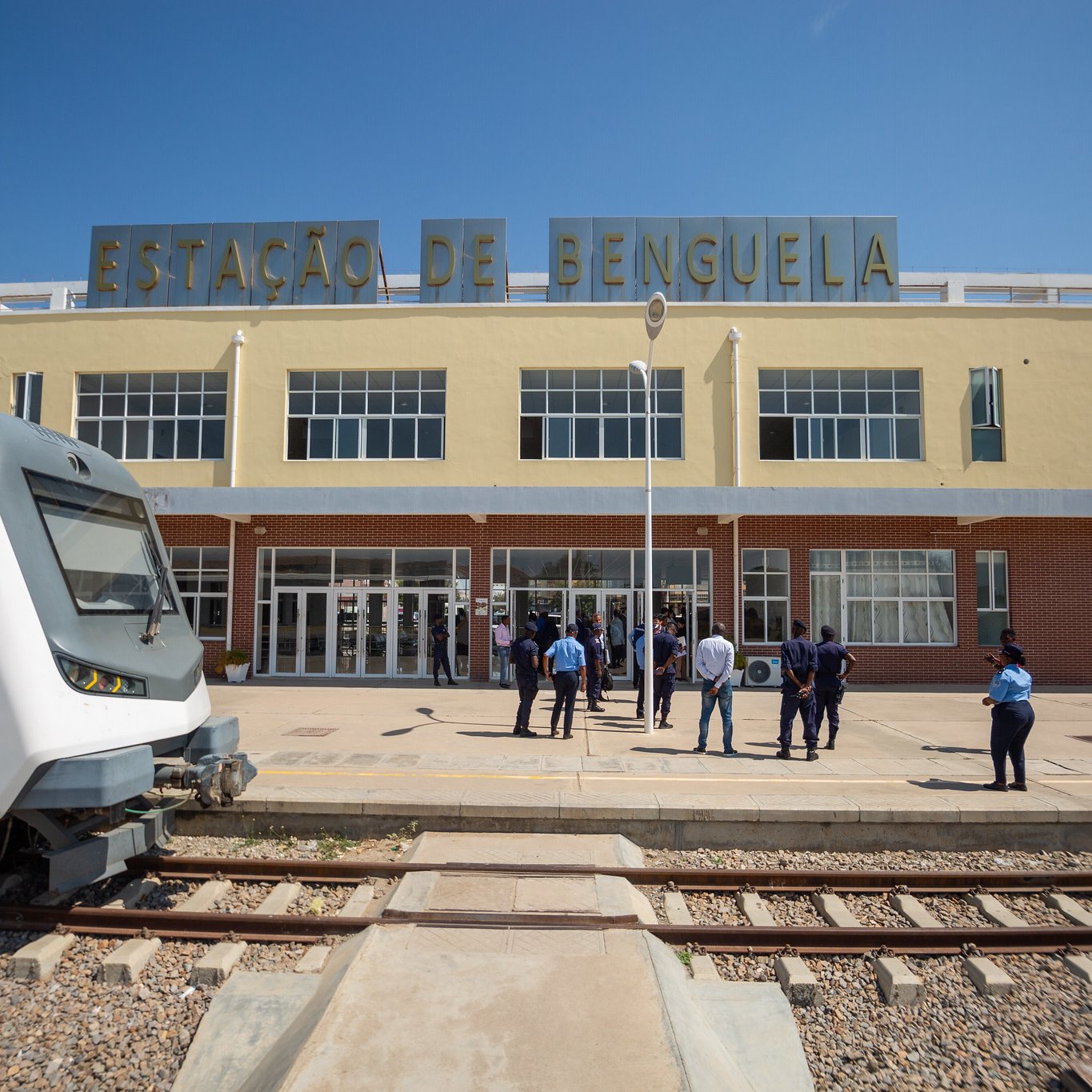 Cette image montre la gare de Benguela, un bâtiment aux couleurs pâles et au style architectural distinctif. À l'avant, on voit un train moderne garé sur une voie ferrée. L'architecture de la gare est caractérisée par des fenêtres spacieuses qui laissent entrer beaucoup de lumière naturelle. Devant la gare, un groupe de personnes, probablement des voyageurs ou du personnel, se tient debout, discutant ou attendant. Le ciel est clair et ensoleillé, créant une ambiance vive et accueillante. Les rails de la gare sont visibles au premier plan, ajoutant au contexte ferroviaire de la scène.