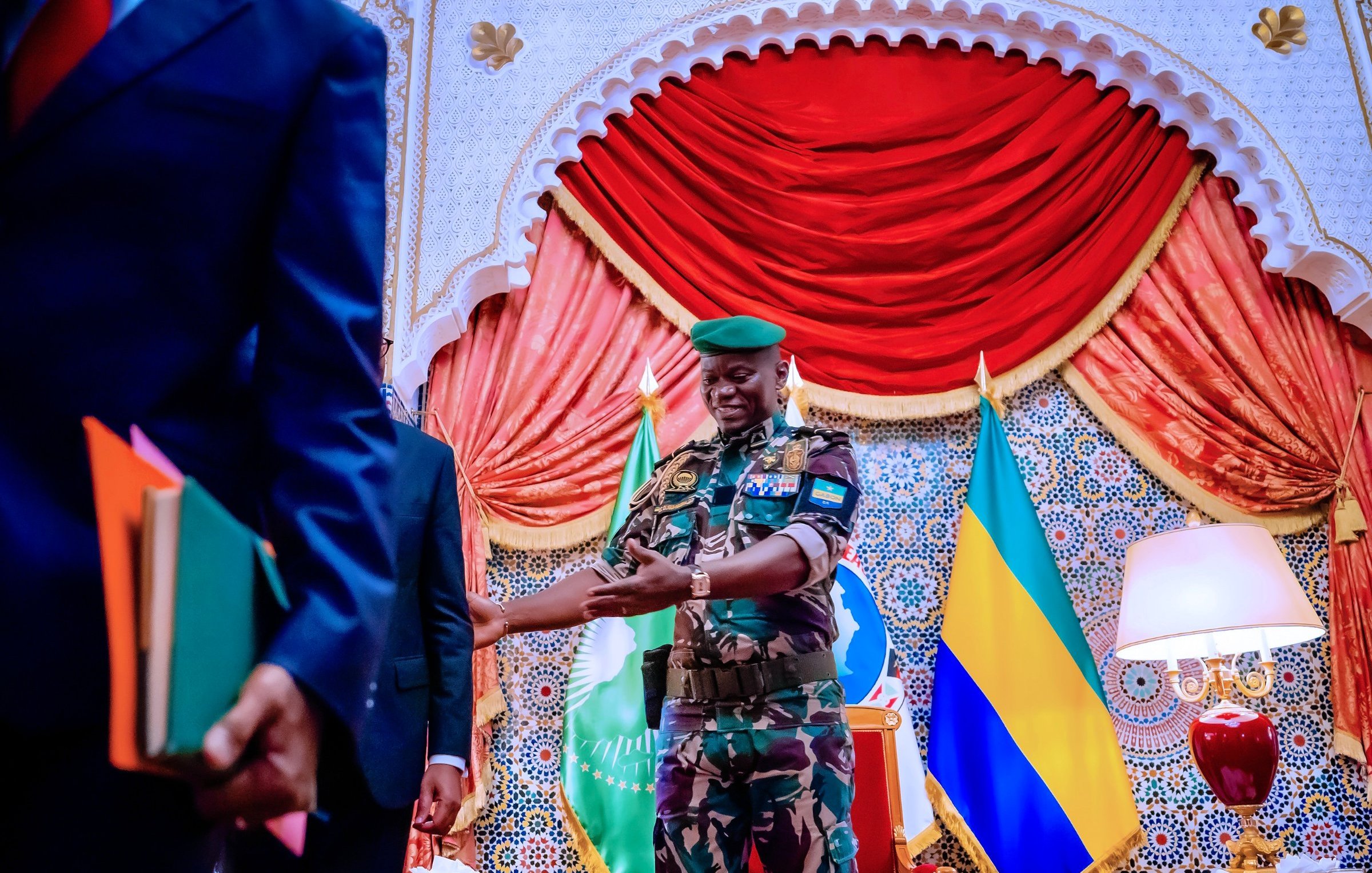 L'image montre un salon d'apparat coloré, avec un décor richement orné et des drapeaux en arrière-plan, dont on distingue le drapeau du Gabon. Au centre, un homme portant un uniforme militaire camouflé et une casquette verte se tient debout avec un sourire accueillant. Il est en train de tendre la main vers une autre personne, probablement pour une poignée de main. Les murs sont décorés de motifs arabes et la scène dégage une atmosphère de formalité et de respect. Une lampe est placée à droite, ajoutant une touche de chaleur à l'environnement.