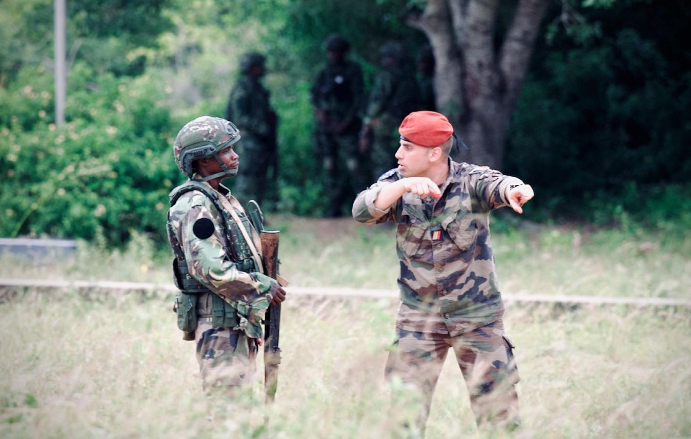 L'image montre deux militaires en discussion dans un environnement extérieur. À gauche, un soldat vêtu d'un uniforme camouflage, portant un casque et tenant un fusil. À droite, un autre militaire, qui porte une tenue camouflage et un béret rouge. Il semble gesticuler en indiquant une direction. En arrière-plan, on aperçoit d'autres soldats flous, ce qui suggère qu'ils sont dans une zone d'entraînement ou opérationnelle, entourée de végétation et d'arbres. L'ambiance est sérieuse et dénote une situation de coordination ou de briefing entre les membres de l'armée.