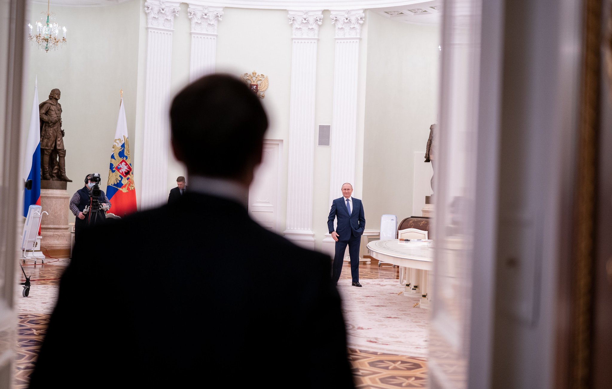 L'image montre une scène dans un intérieur officiel, probablement un palais ou un bureau gouvernemental. Au premier plan, on voit le dos d'une personne en costume sombre, qui semble faire face à une autre personne située à une certaine distance. Cette deuxième personne, qui est clairement identifiable comme une figure politique importante, se tient debout dans une pièce au décor élégant, avec une lumière douce. À l'arrière-plan, on peut distinguer des drapeaux russe et peut-être d'autres éléments symboliques. Le sol est en marbre, et les murs sont peints dans des teintes claires, ajoutant une ambiance solennelle à l'ensemble. L'atmosphère de l'image suggère un moment de rencontre ou de dialogue important.