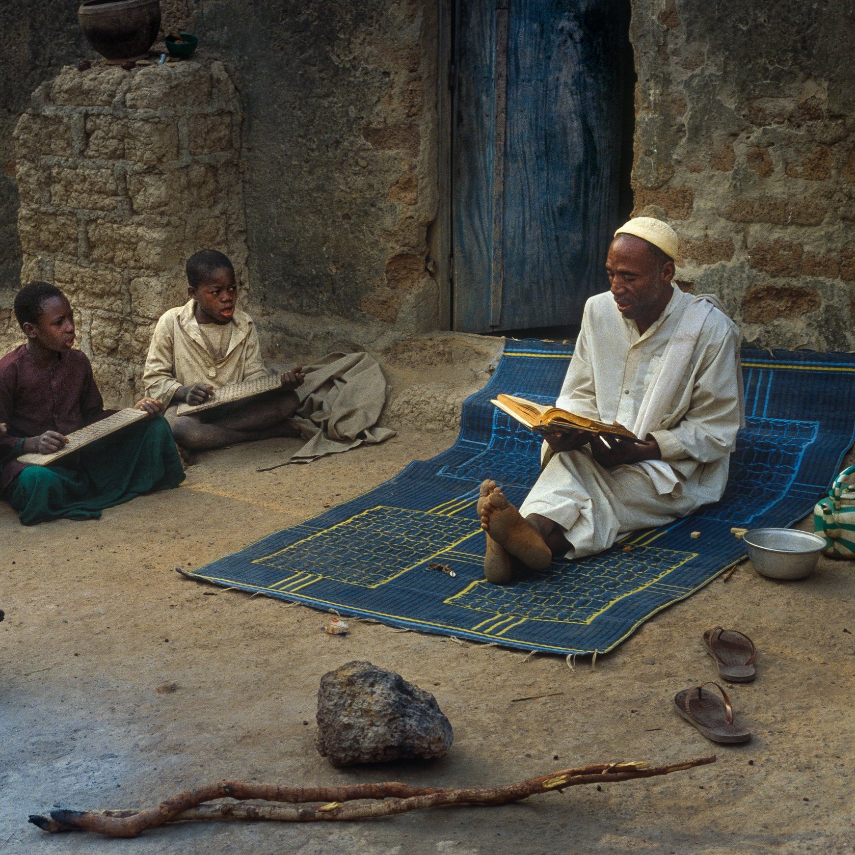 L'image montre un enseignant assis sur un tapis à l'extérieur d'une maison en terre. Il lit un livre avec attention, tandis que plusieurs enfants sont rassemblés autour de lui, concentrés sur des planches ou des livres qu'ils tiennent. L'ambiance est calme et studieuse. On peut aussi voir un poulet se déplacer au sol, ajoutant une touche de vie rurale à la scène. Les murs de la maison sont simples, et la lumière naturelle éclaire l'espace, créant une atmosphère chaleureuse et conviviale.